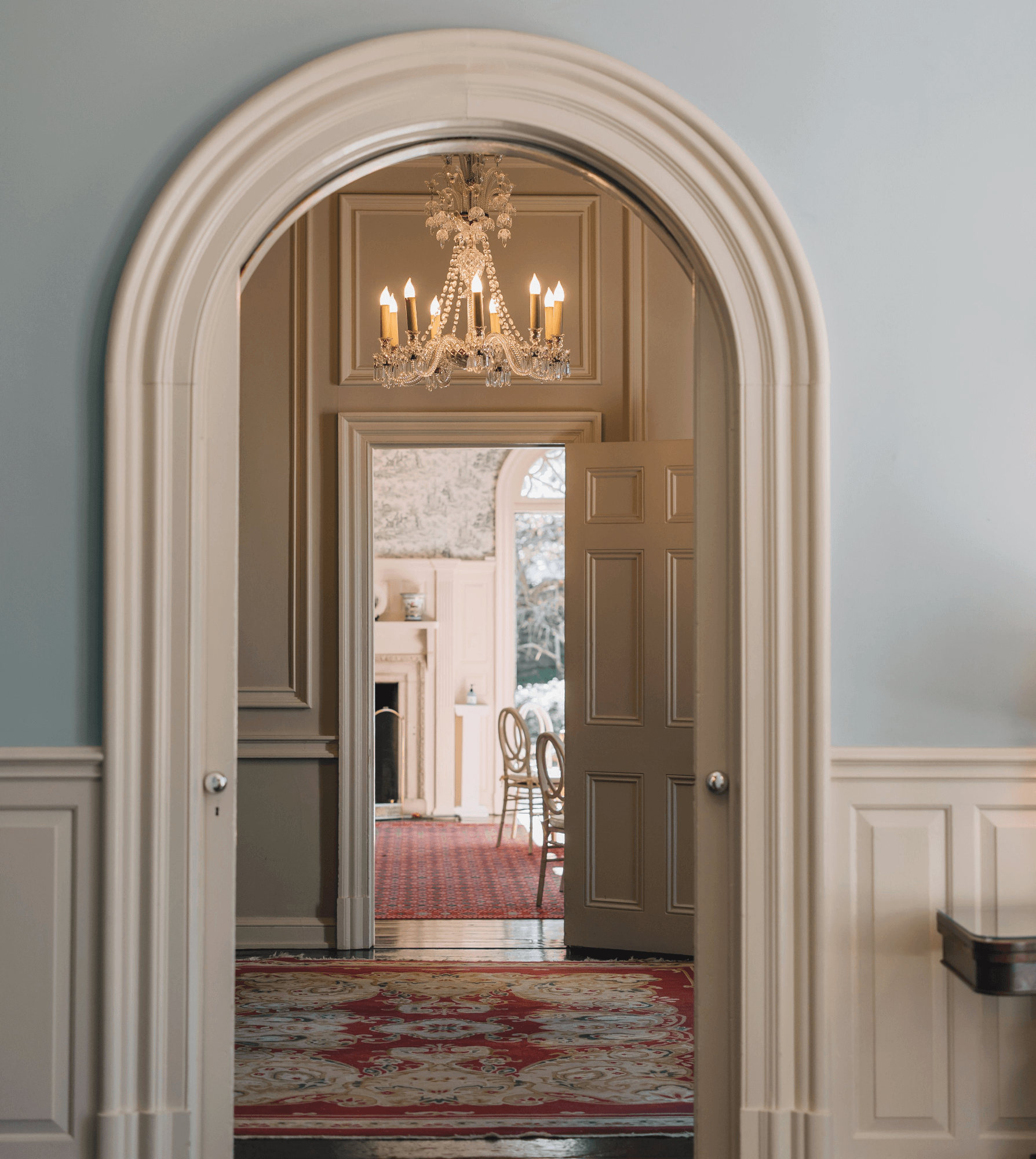 A hallway with a white arched doorway leads to a room with an open door, chandelier, and chairs. An ornate rug covers the floor.