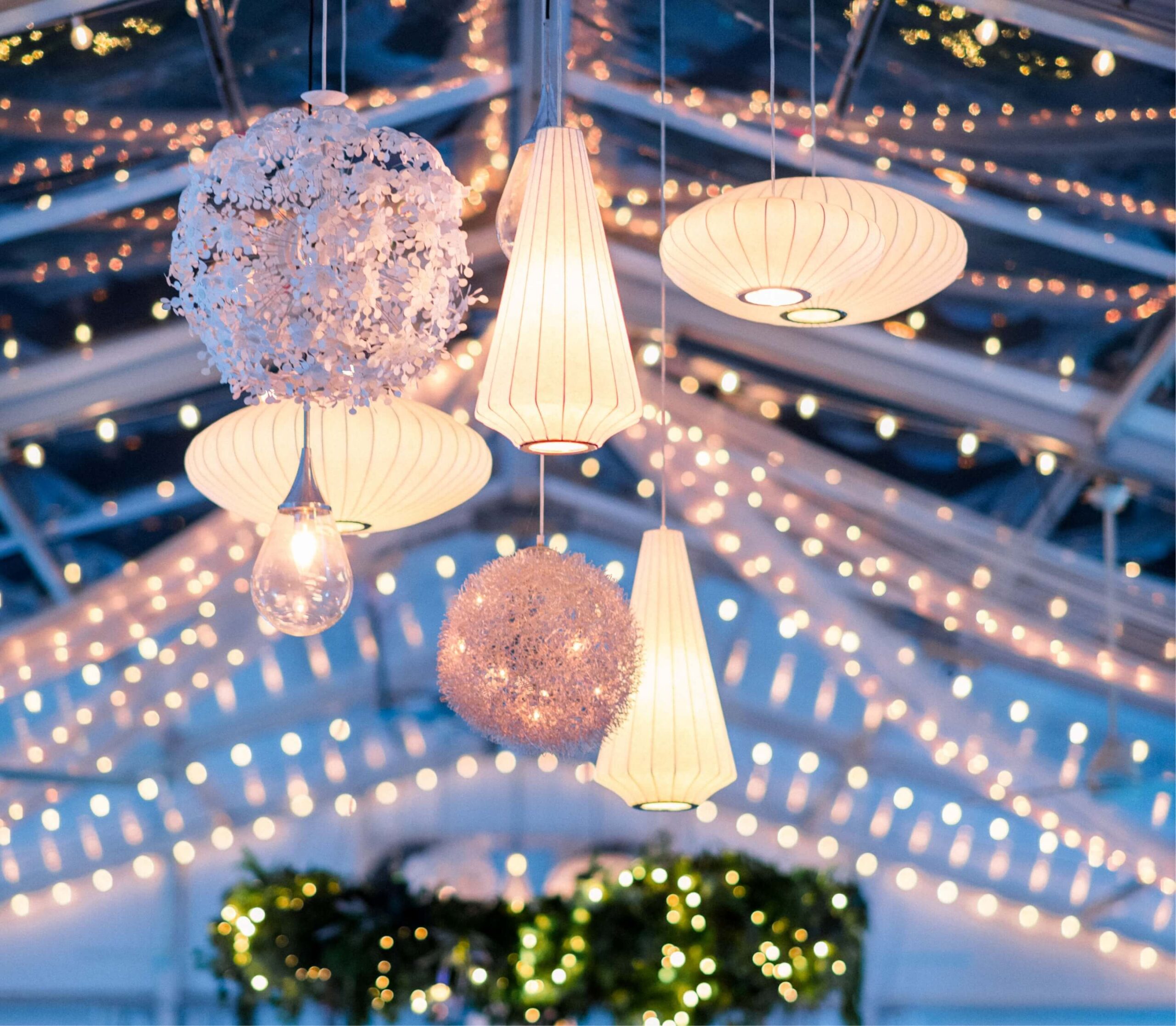 Hanging decorative lights including lanterns and floral designs underneath a clear roof, surrounded by string lights with a blurred bokeh effect.