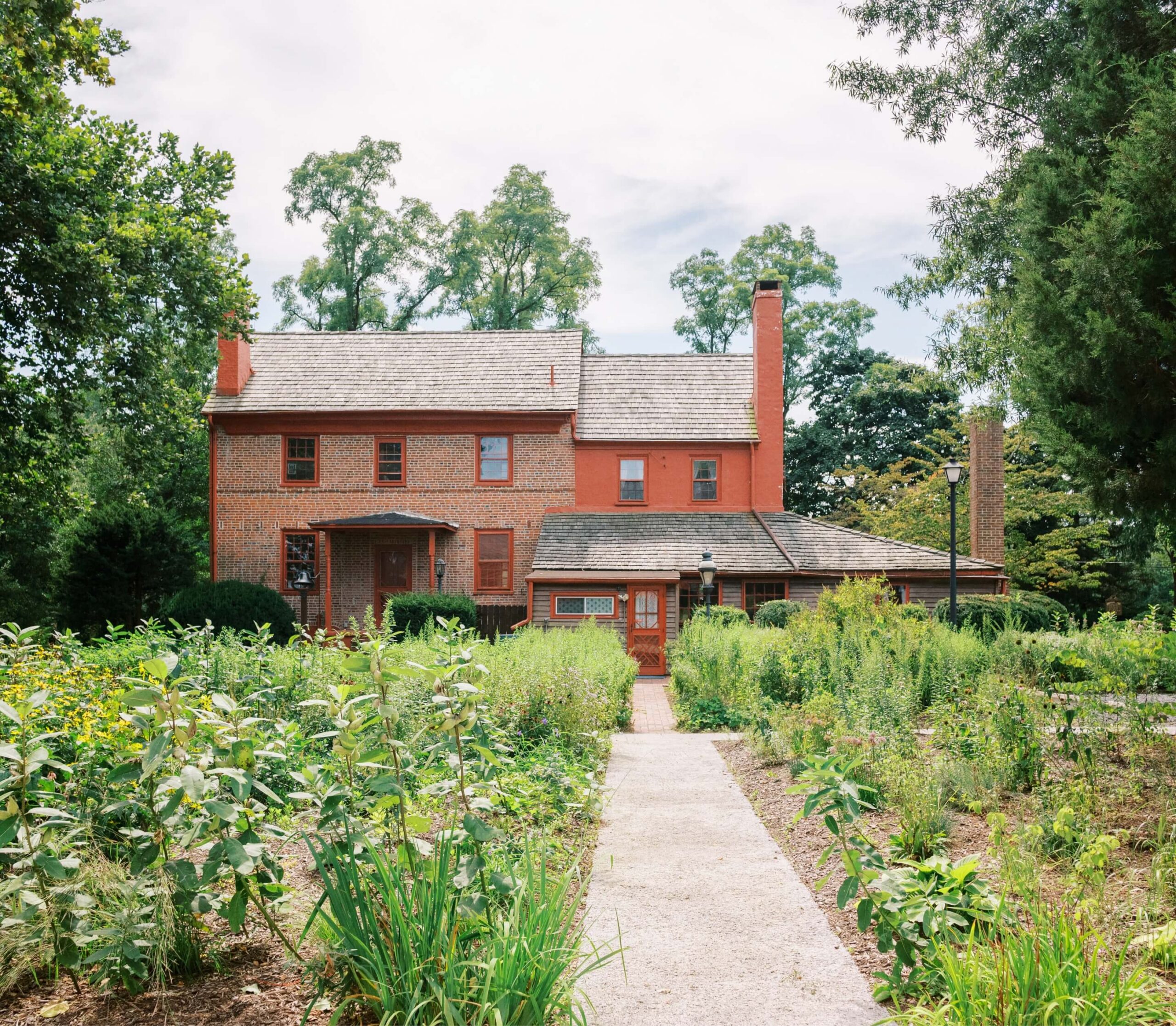Two-story red brick house with a wooden shingle roof, surrounded by greenery and plants, with a stone pathway leading to the entrance.
