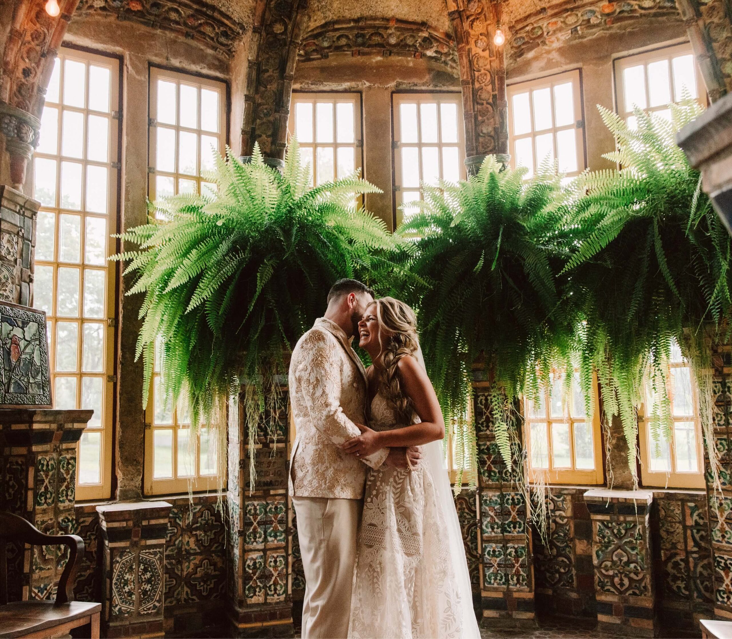Bride and groom embrace in an ornate room with large windows and hanging ferns in the background.