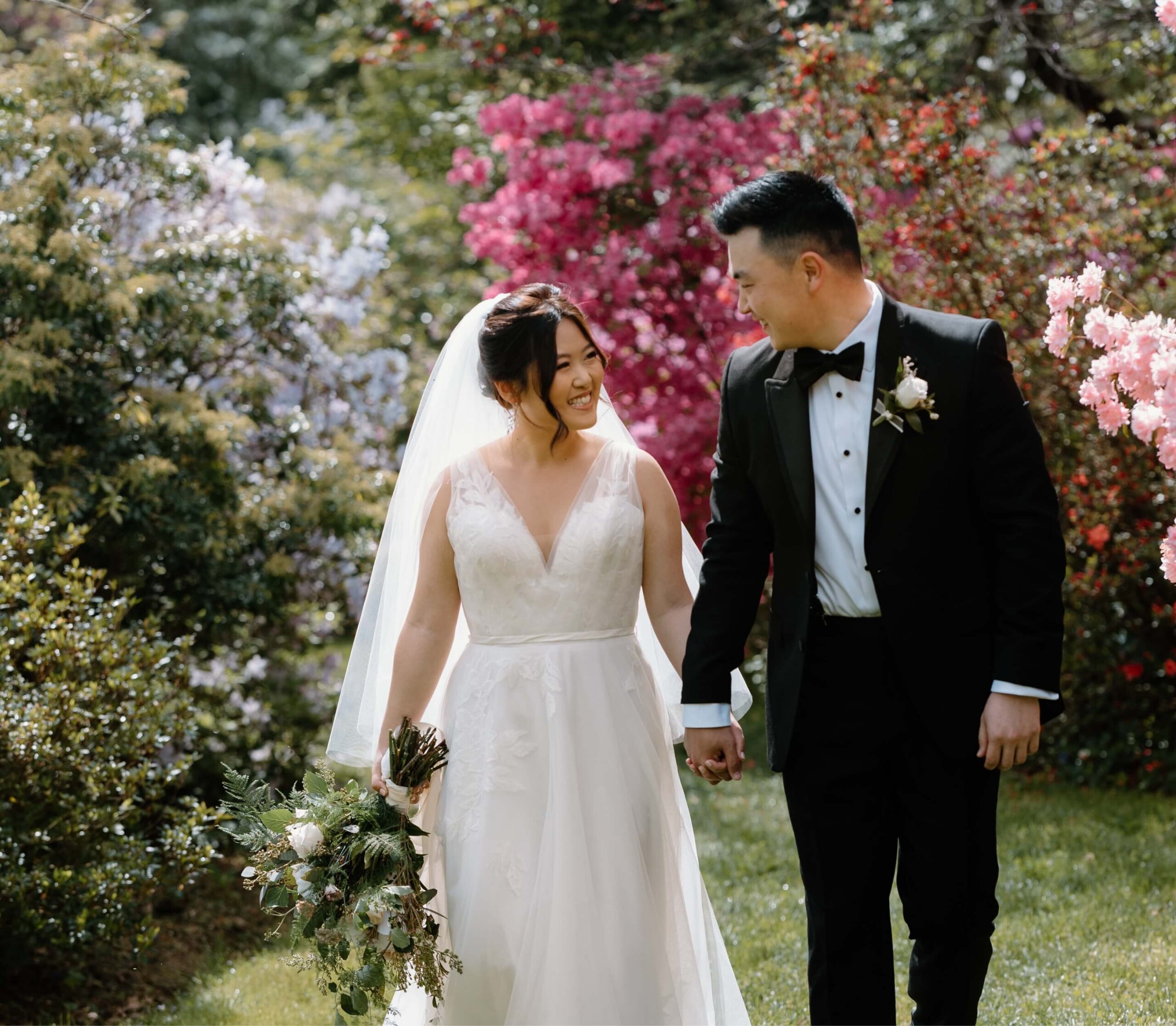 A bride and groom smile at each other while walking hand in hand through a garden with vibrant flowers. The bride holds a bouquet and wears a veil; the groom is in a black tuxedo.