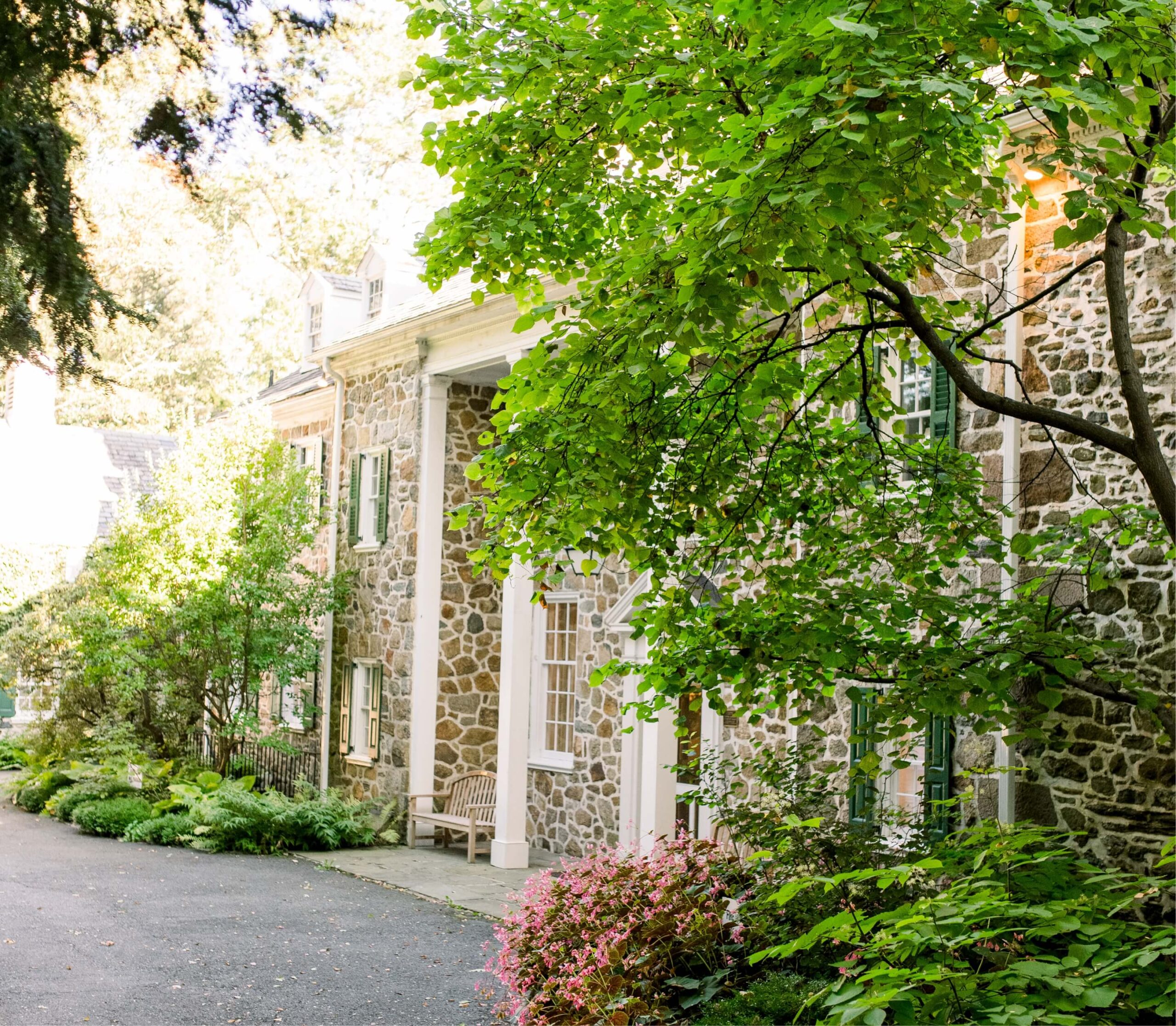Historic stone house with green shutters, surrounded by lush greenery and colorful flowers, under a bright, sunny sky.