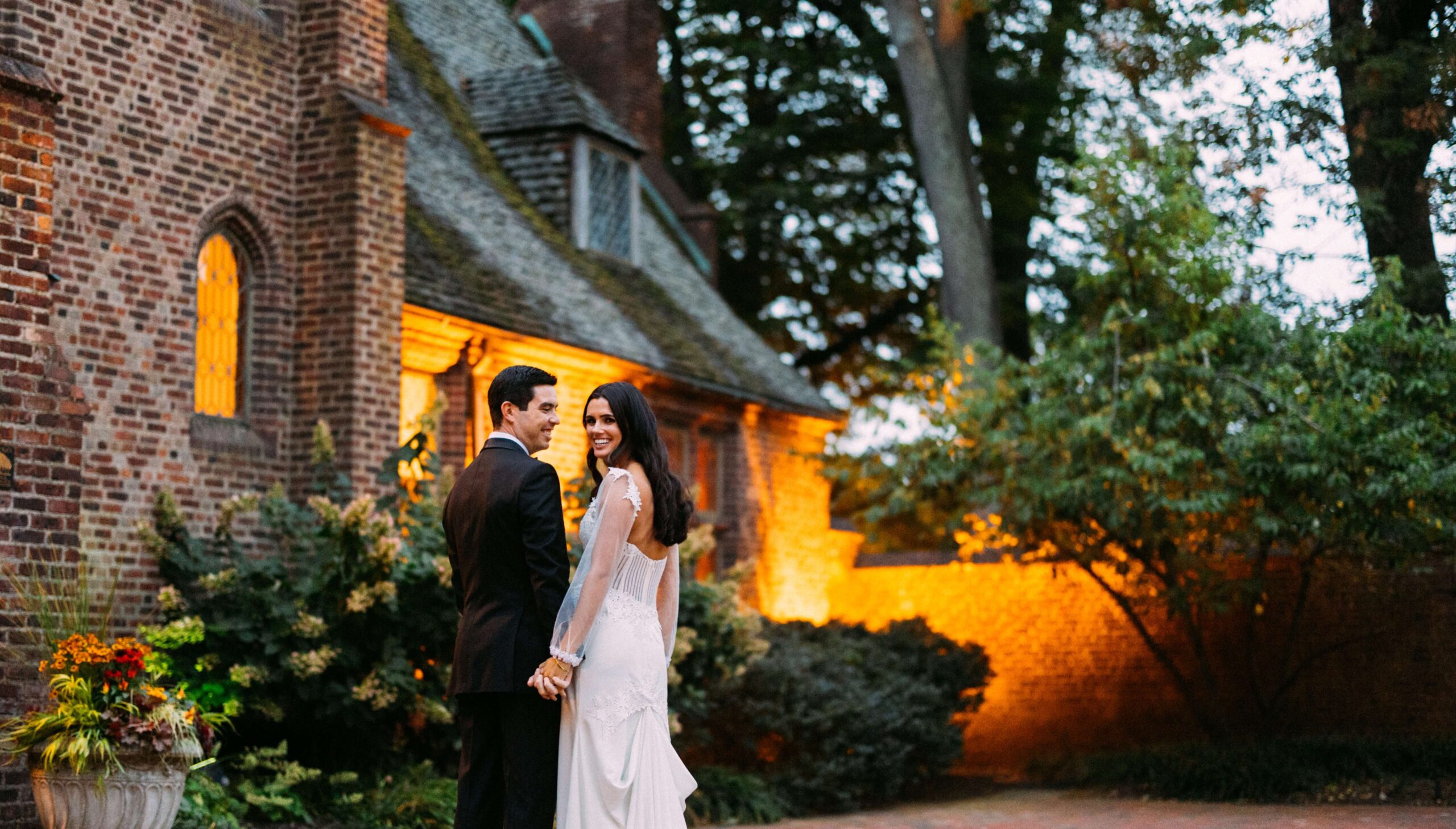 A couple in formal attire stands holding hands in front of a brick building surrounded by trees and lit by warm lights.