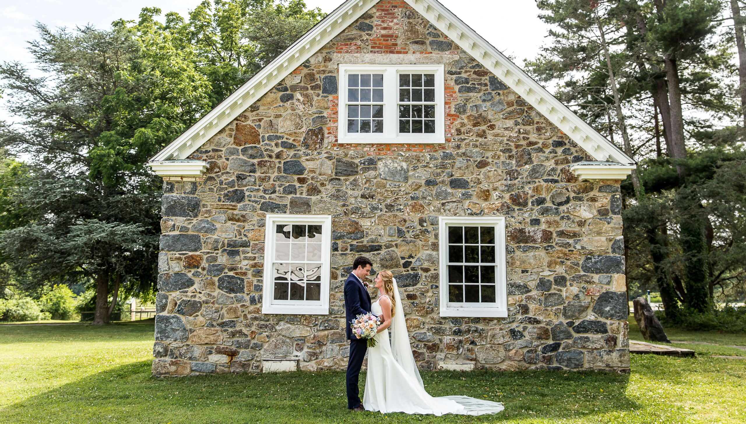 A bride and groom stand facing each other in front of a stone building on a sunny day, surrounded by green grass and trees.
