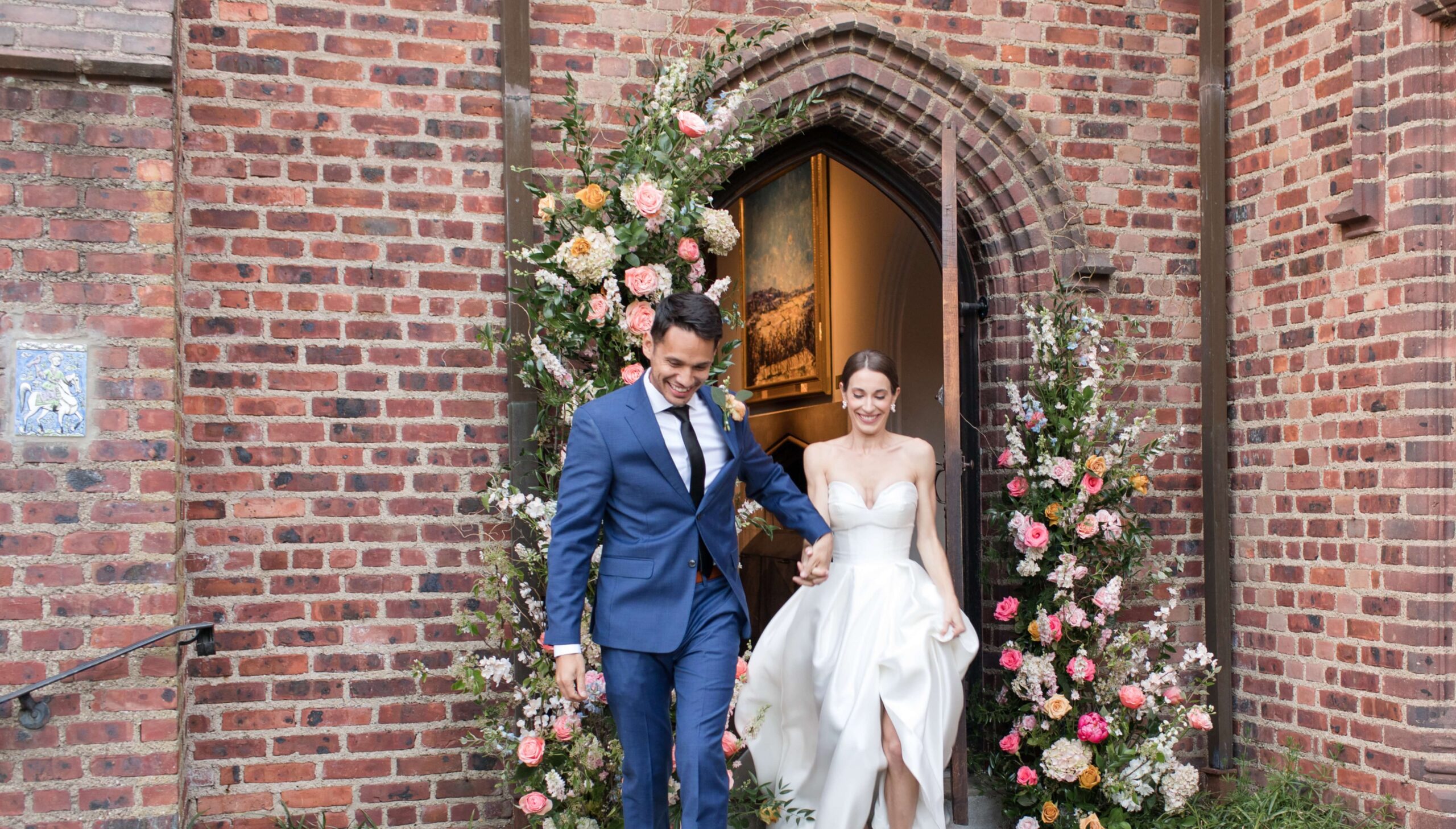 A couple in formal attire exits a brick building through an archway decorated with flowers.