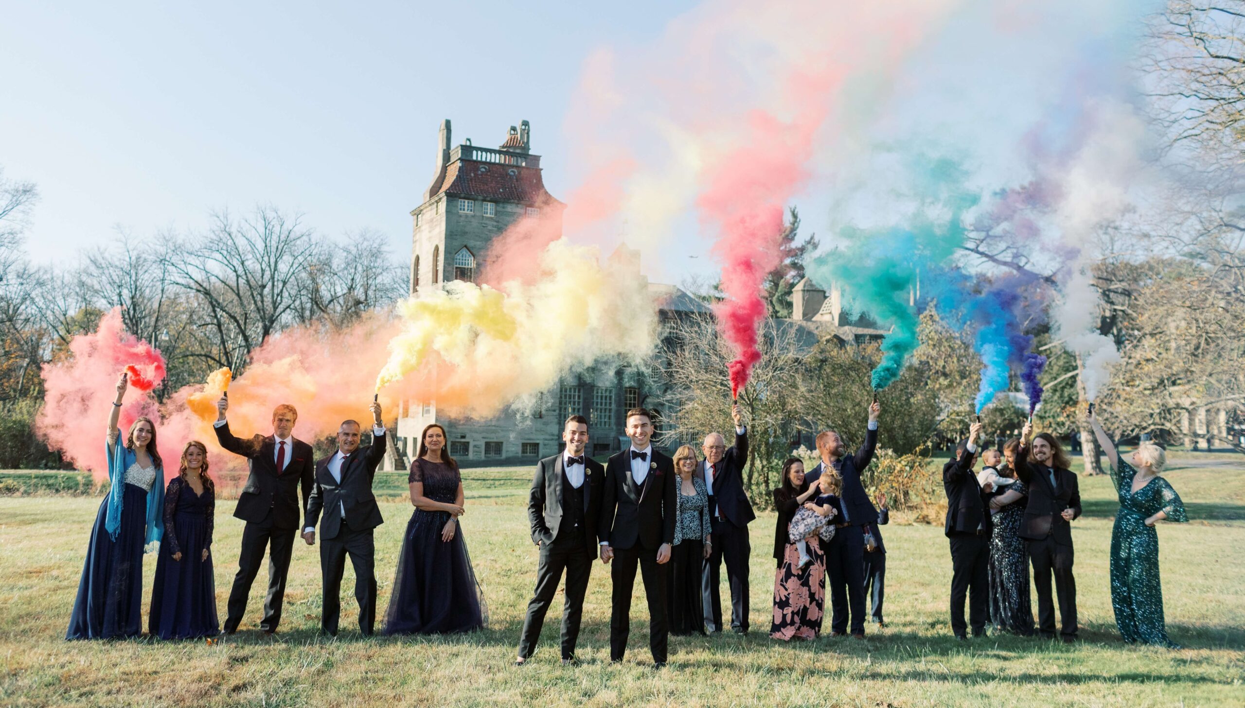 A group of people in formal attire stand in a field, holding colorful smoke flares in shades of yellow, red, blue, purple, and green, with a large building in the background.