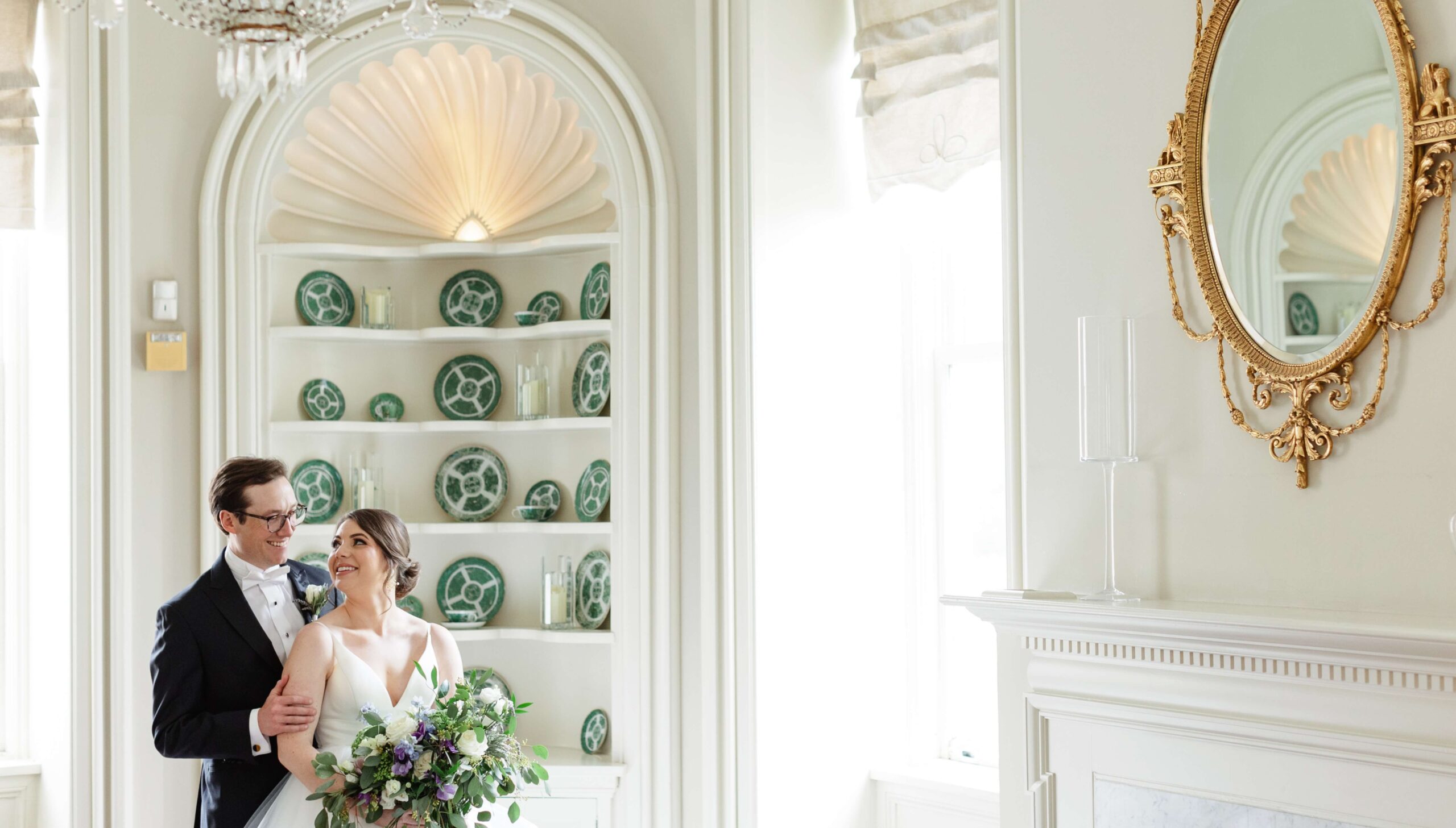 A couple in wedding attire stands smiling in an elegant white room with green decorative plates on shelves and a gold-framed mirror on the wall.