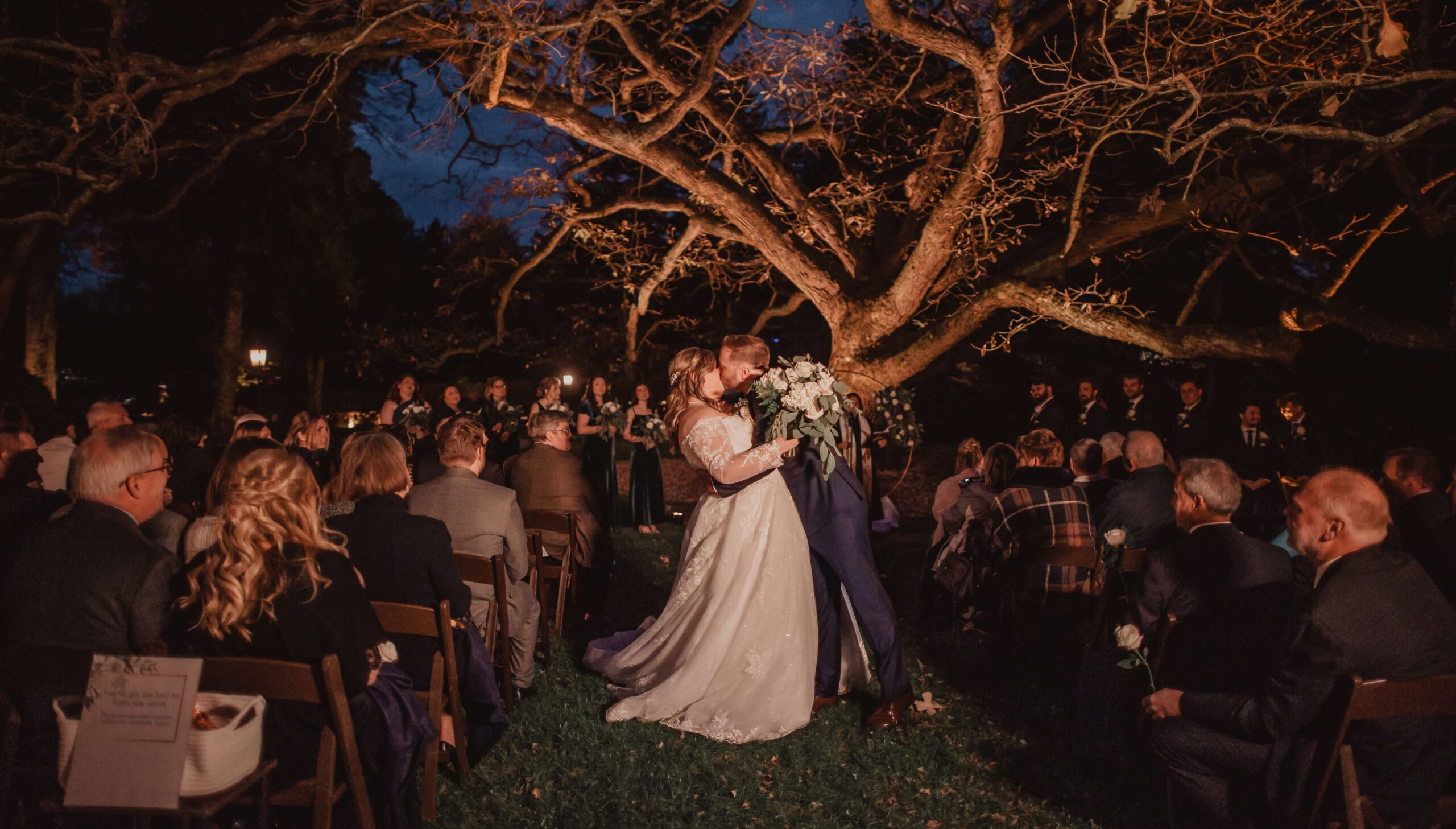A couple kisses under a large, illuminated tree during an outdoor nighttime wedding ceremony, surrounded by seated guests.