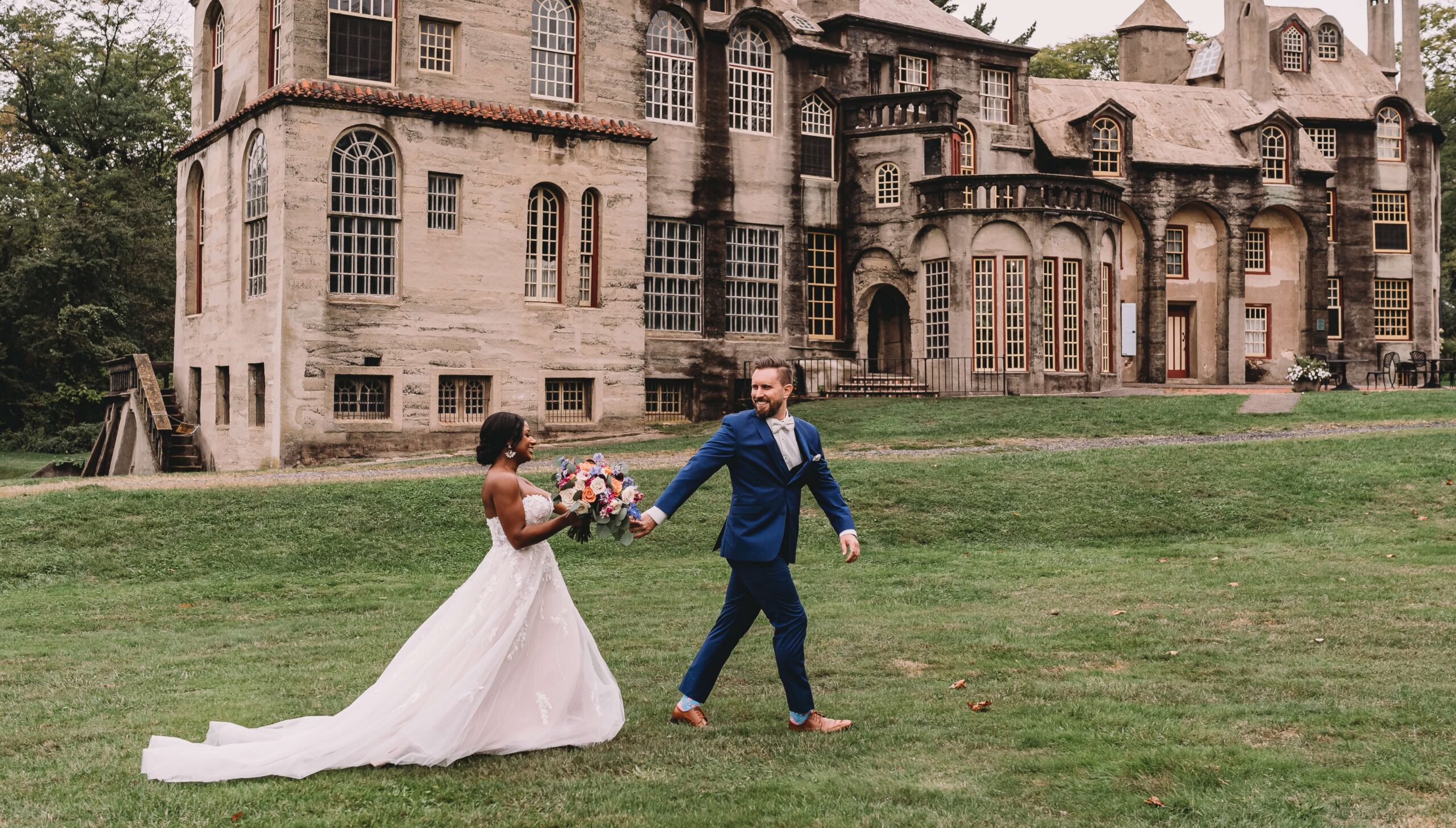 A couple in wedding attire walks hand in hand across a grassy area in front of a large, historic building.