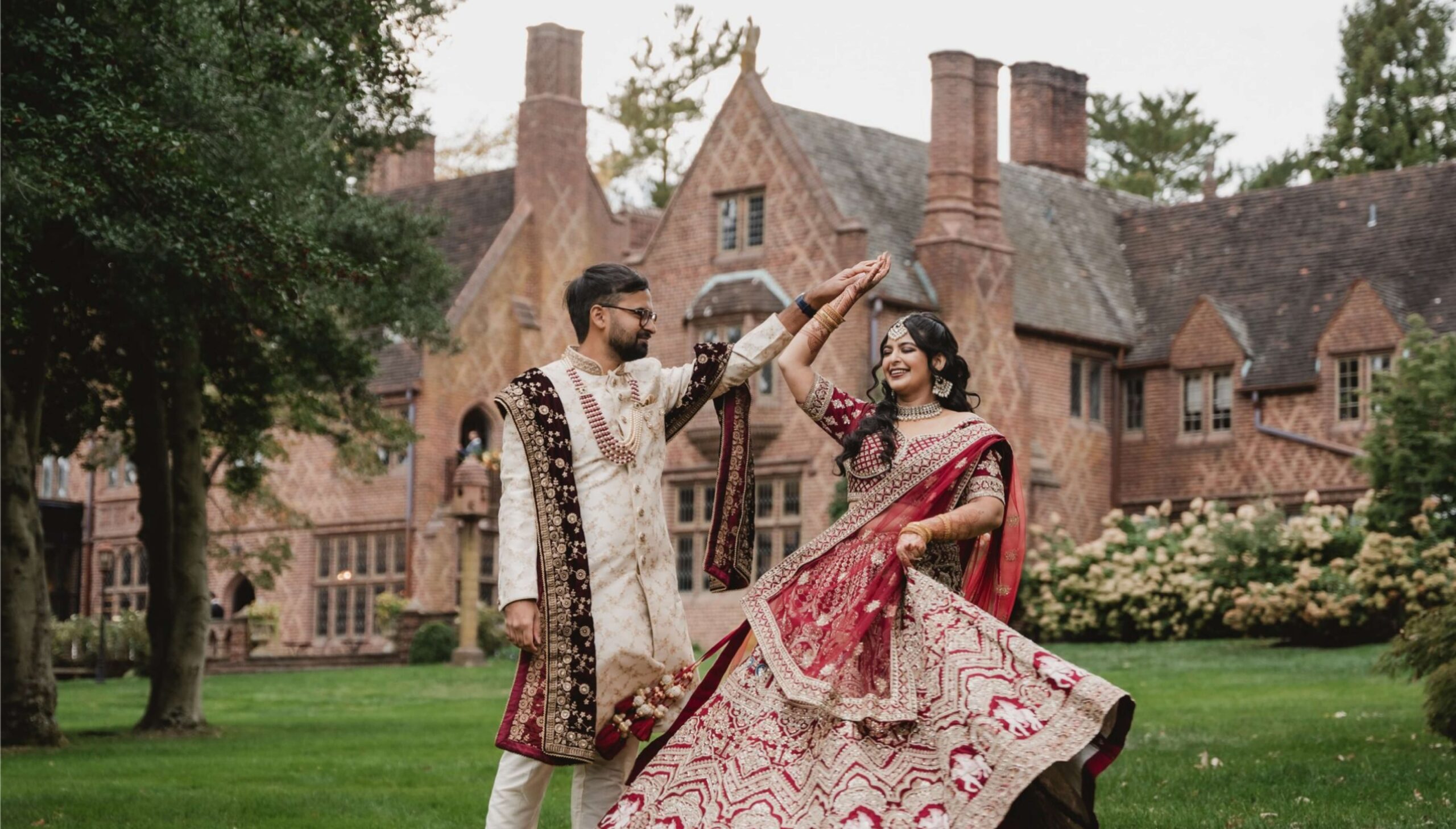 Couple in traditional Indian attire dancing on a grassy lawn in front of a large, brick mansion.