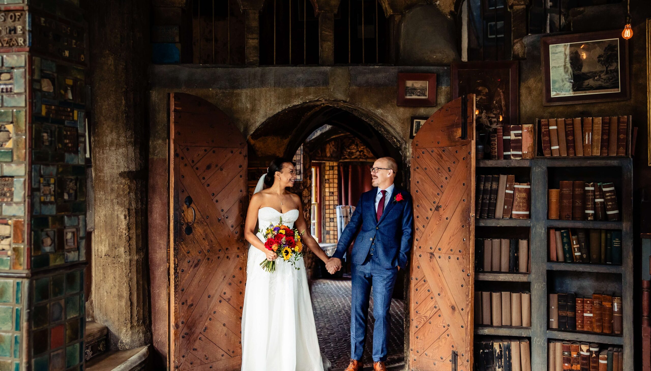 A bride and groom hold hands, smiling at each other, standing in a room with wooden doors and shelves filled with books.
