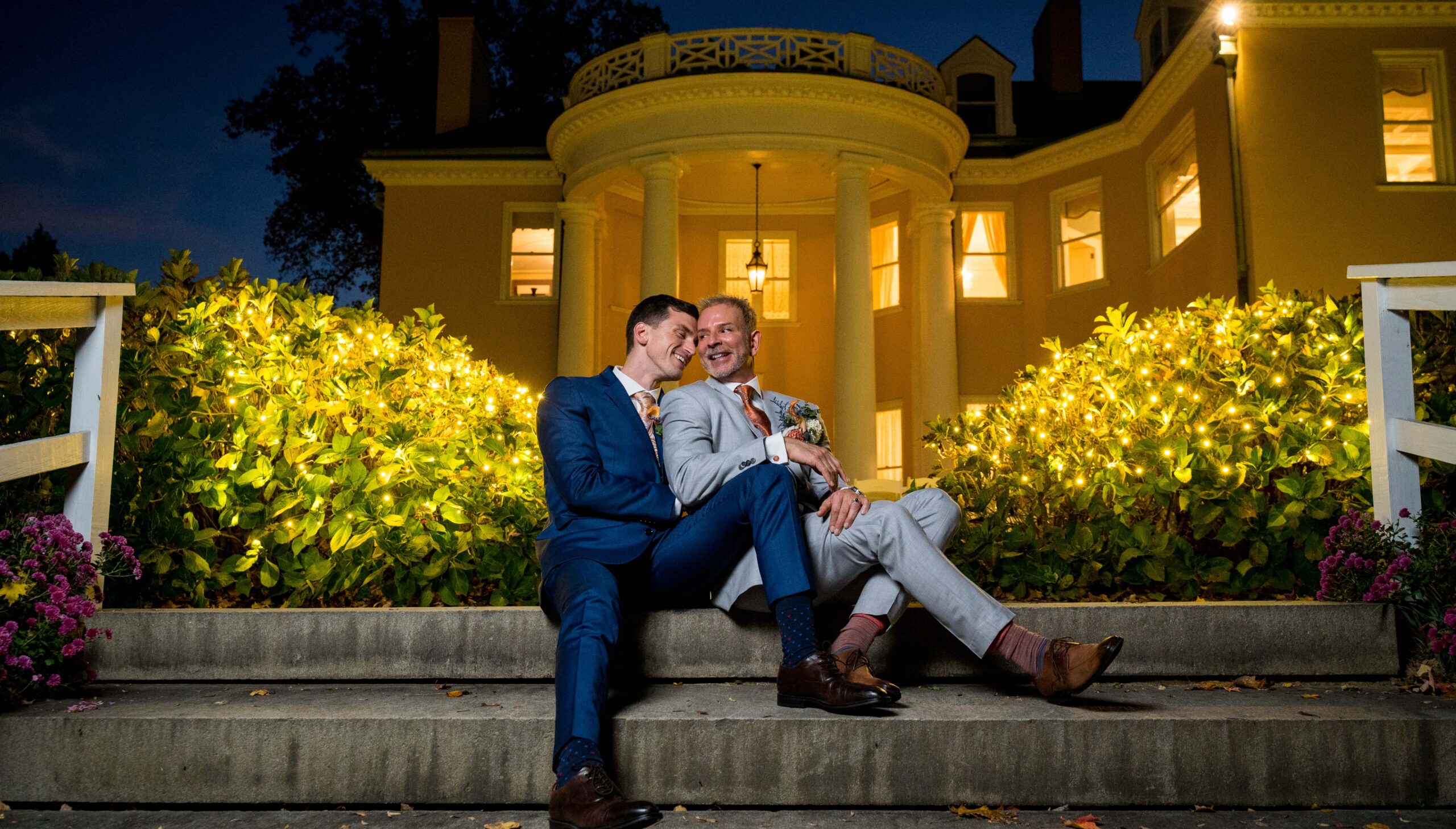 Two men in suits sit closely on steps outside a lit-up building at night, surrounded by illuminated shrubs.