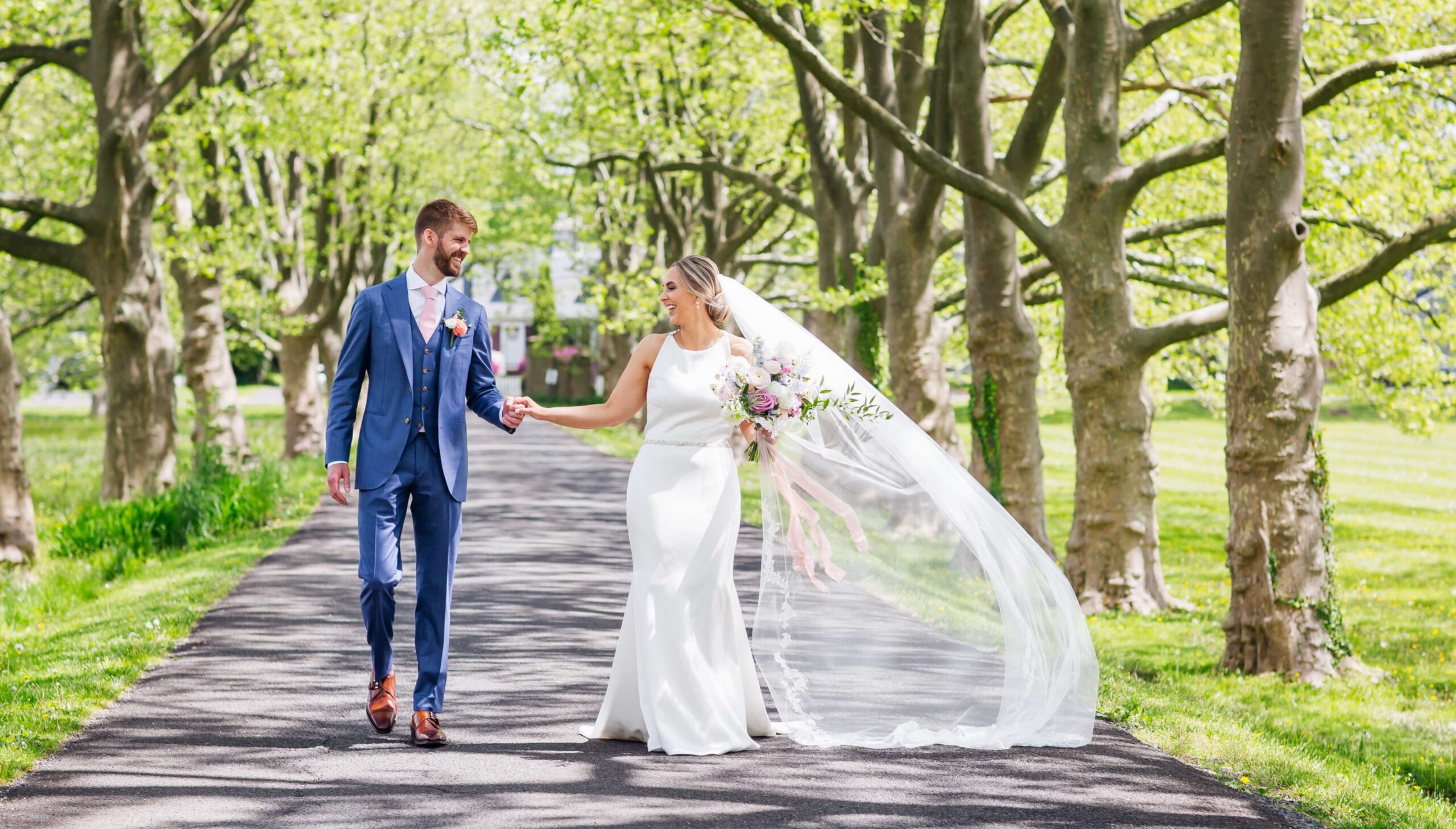 A bride and groom walk hand in hand on a tree-lined path. The bride wears a long white dress with a veil, and the groom is in a blue suit. They both smile at each other.