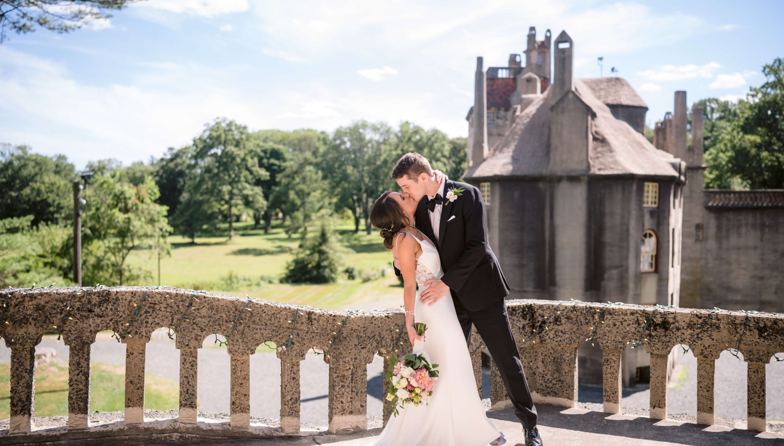 A bride and groom share a kiss on a stone balcony with a historic building and lush greenery in the background.