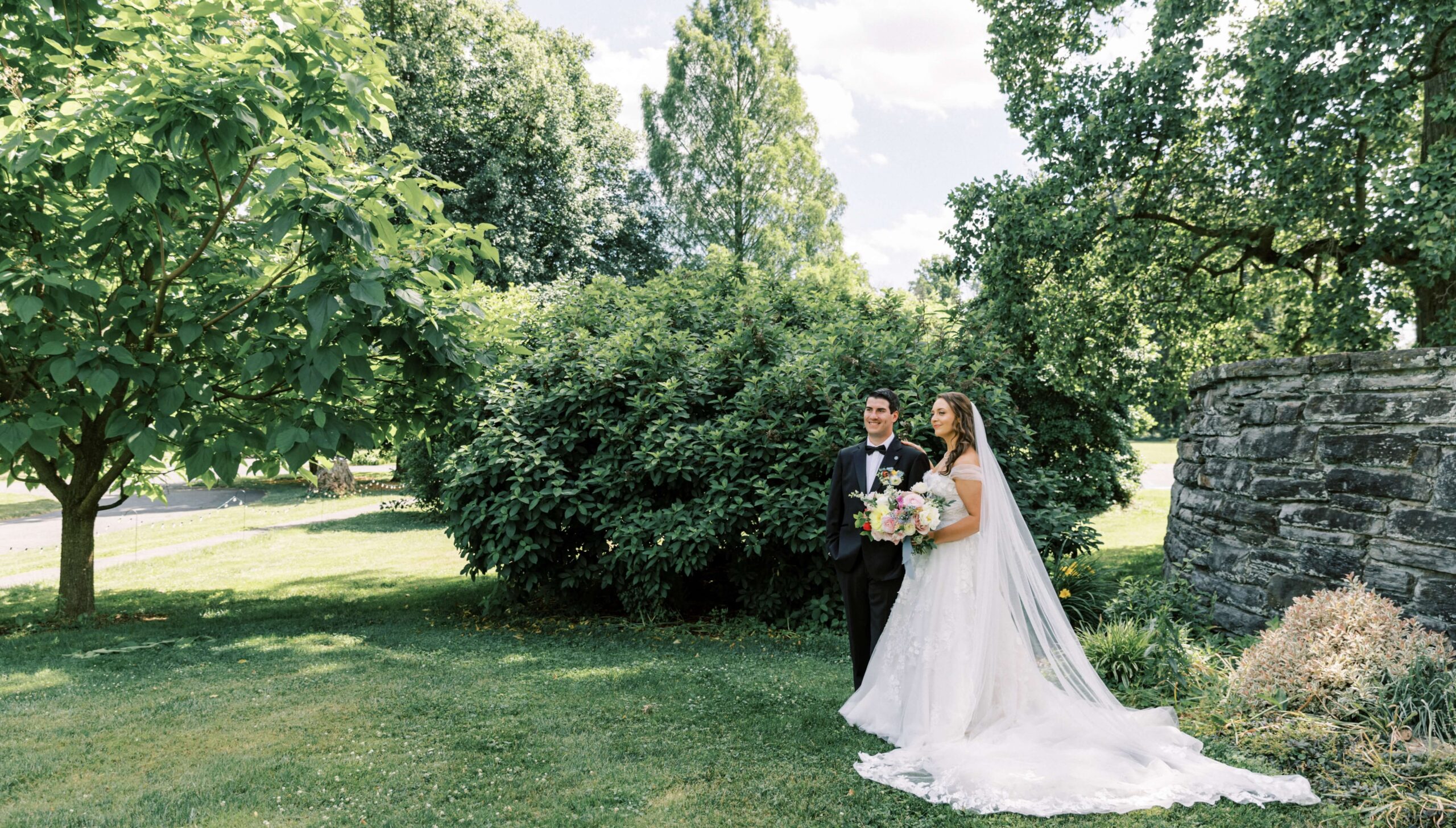 A bride and groom stand together outdoors on grass, surrounded by greenery. The bride holds a bouquet and wears a long veil.