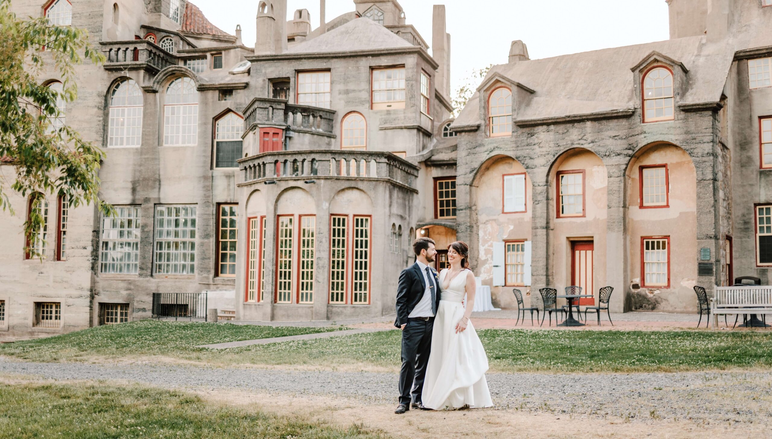 A couple in formal attire stands in front of a large, historic stone building with arched windows and red trim.