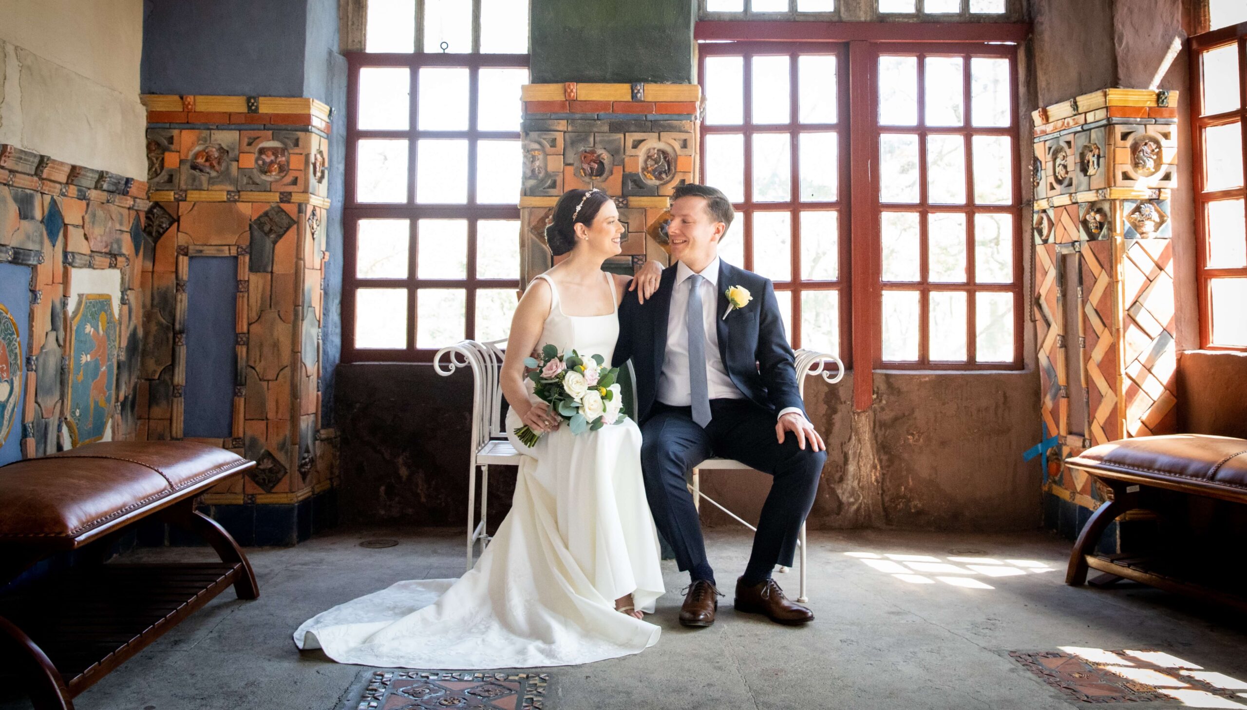A bride and groom sit on an indoor bench, smiling at each other. The room features large windows and decorative tiles.