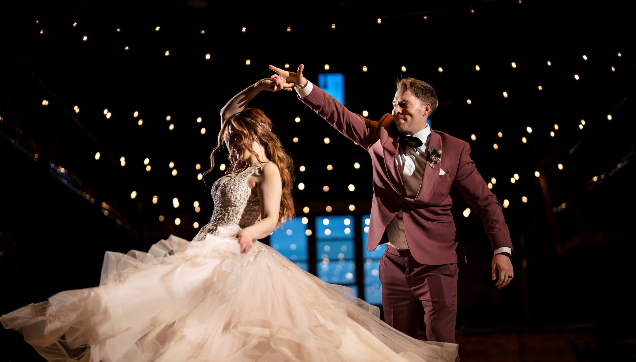 A bride and groom dance under string lights. The bride's gown flares as she twirls, and the groom holds her hand, smiling.
