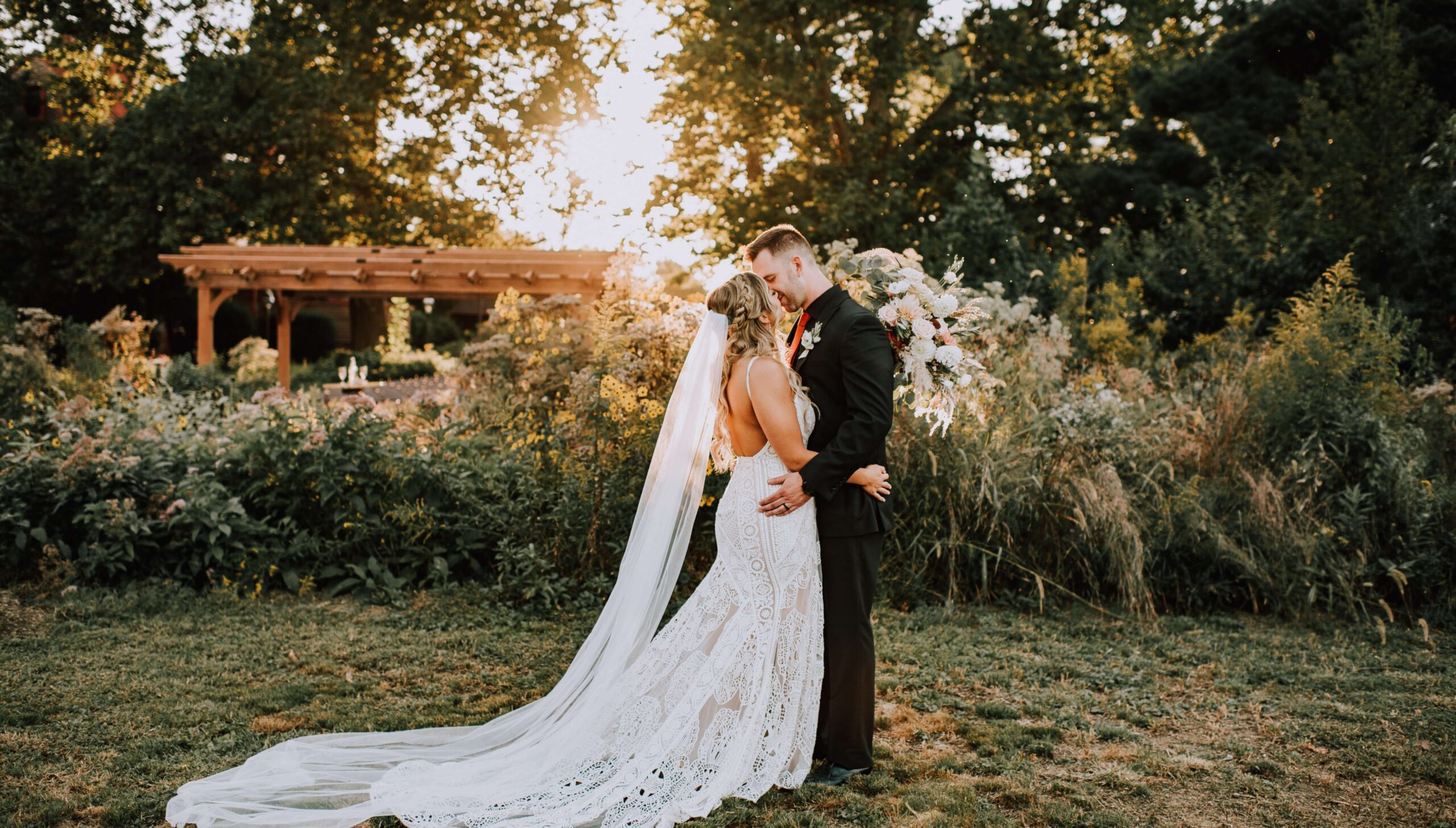 Bride and groom standing in a garden, embracing under a wooden pergola. She wears a long veil and lace gown, and he is in a dark suit. The sun sets in the background.