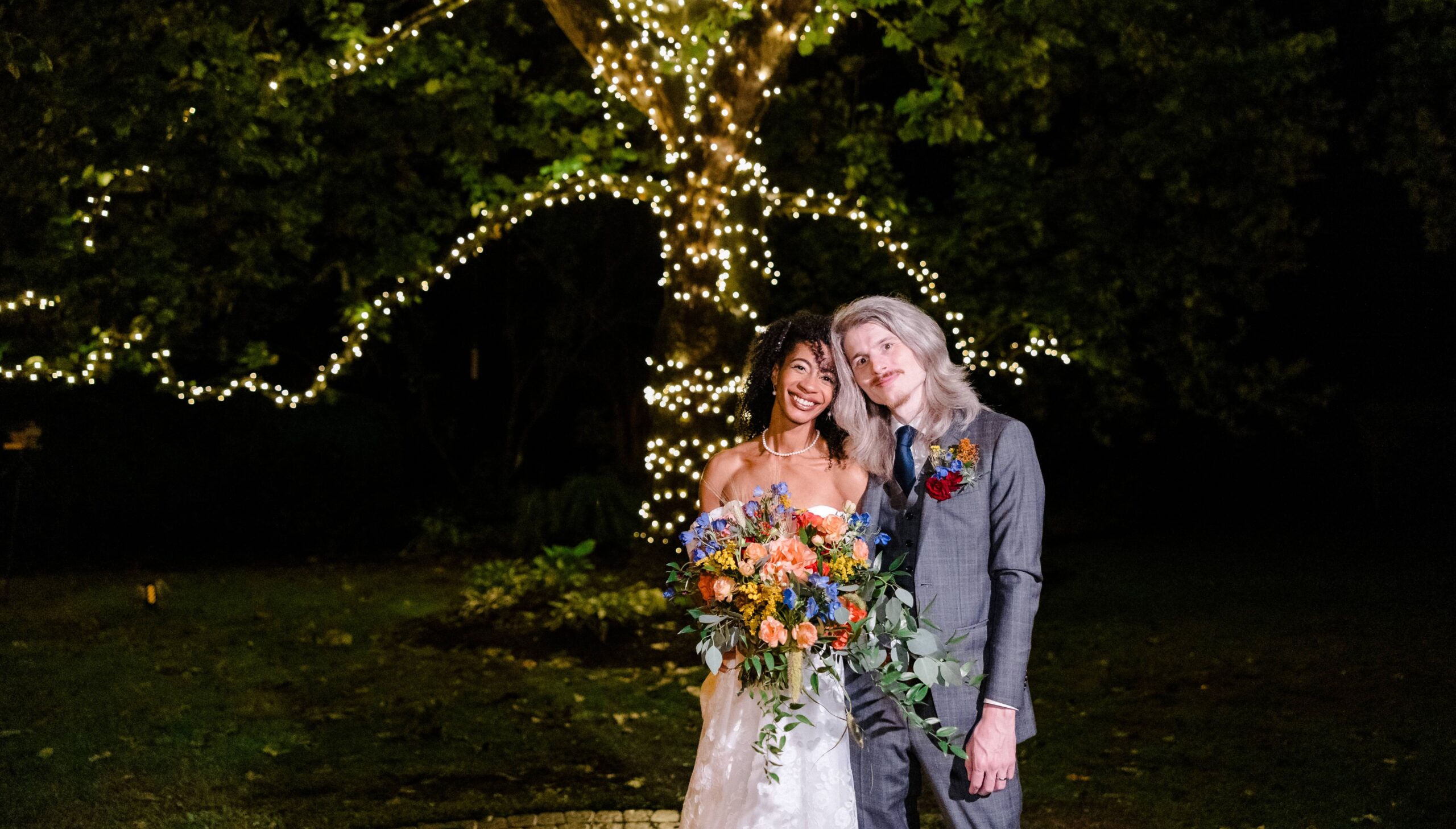 Bride and groom stand together at night with a bouquet, with a tree wrapped in string lights in the background.
