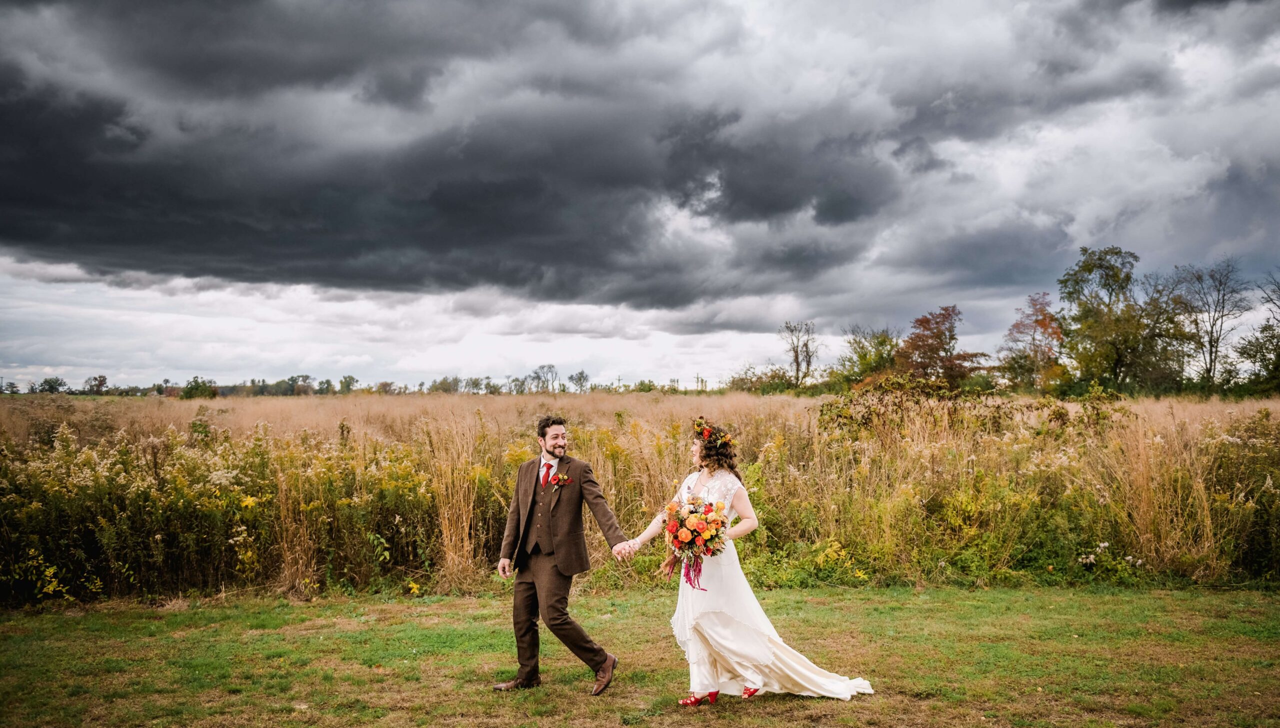 A couple in wedding attire walks hand in hand through a field under a dramatic cloudy sky.