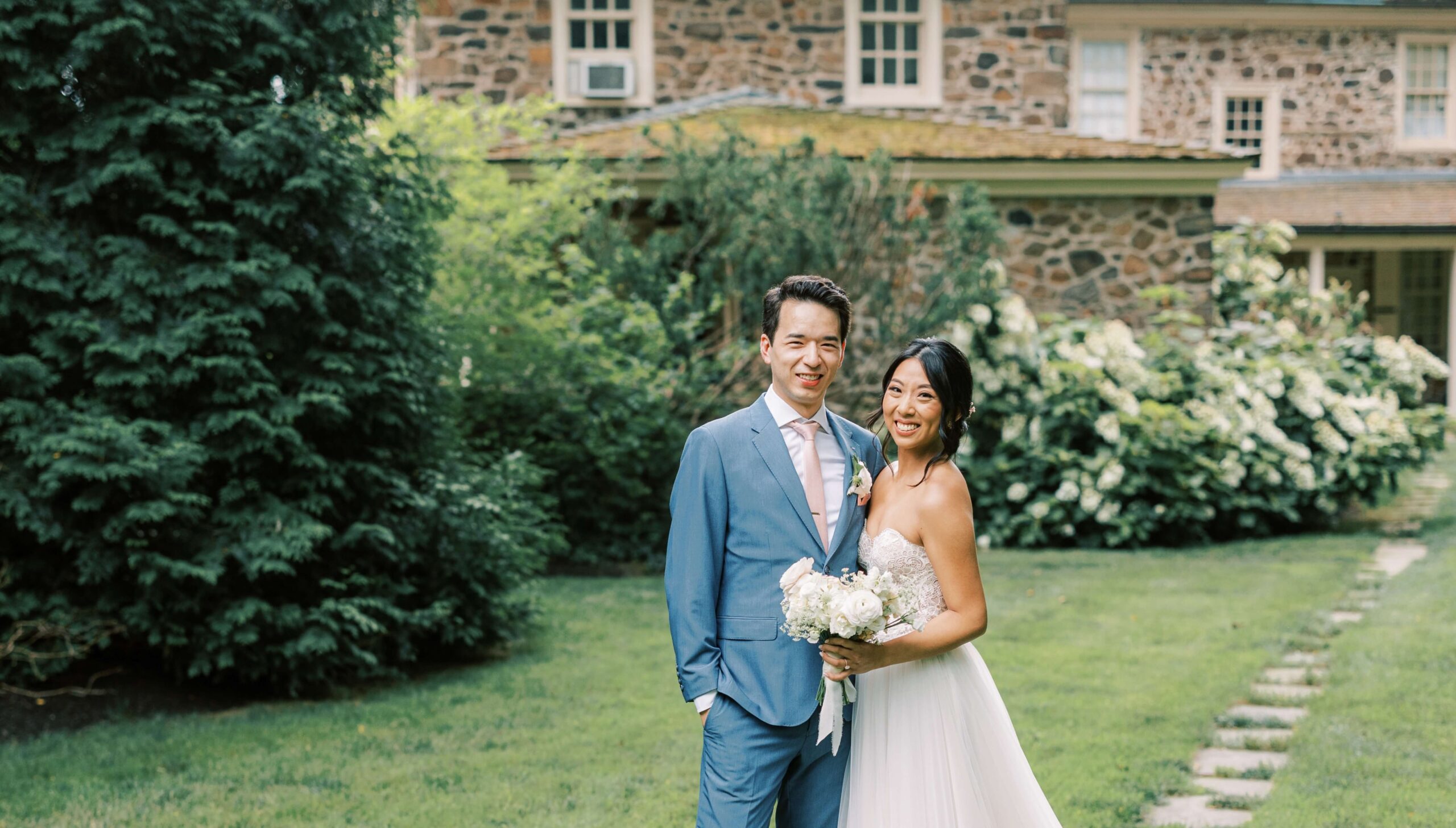 A couple stands outdoors on grass in formal attire; the man in a suit and the woman in a wedding dress holding a bouquet, with a stone house and greenery in the background.
