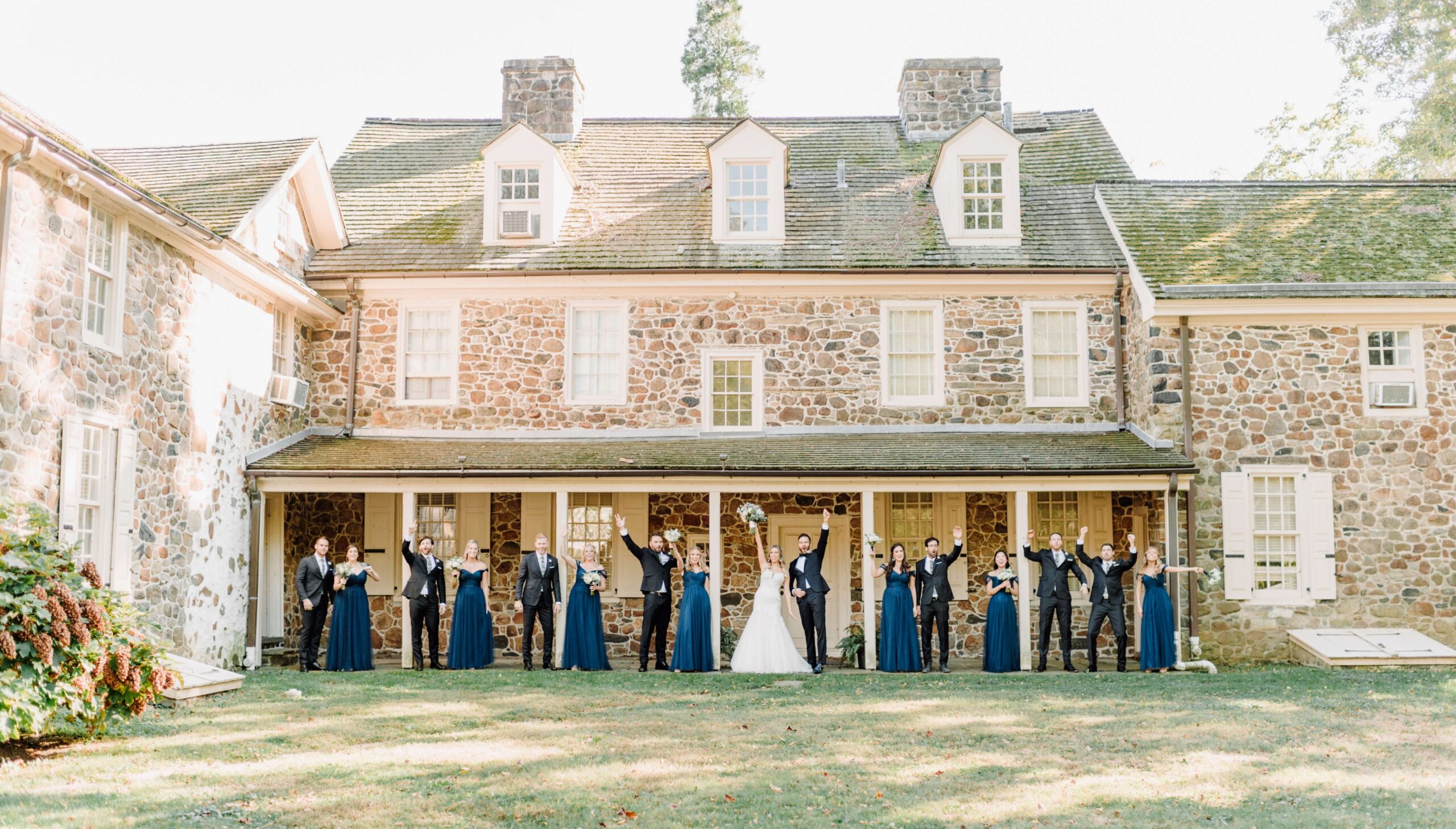 A wedding party poses on the lawn in front of a large stone house, with the bride and groom at the center and bridesmaids and groomsmen in dark formal wear on either side.