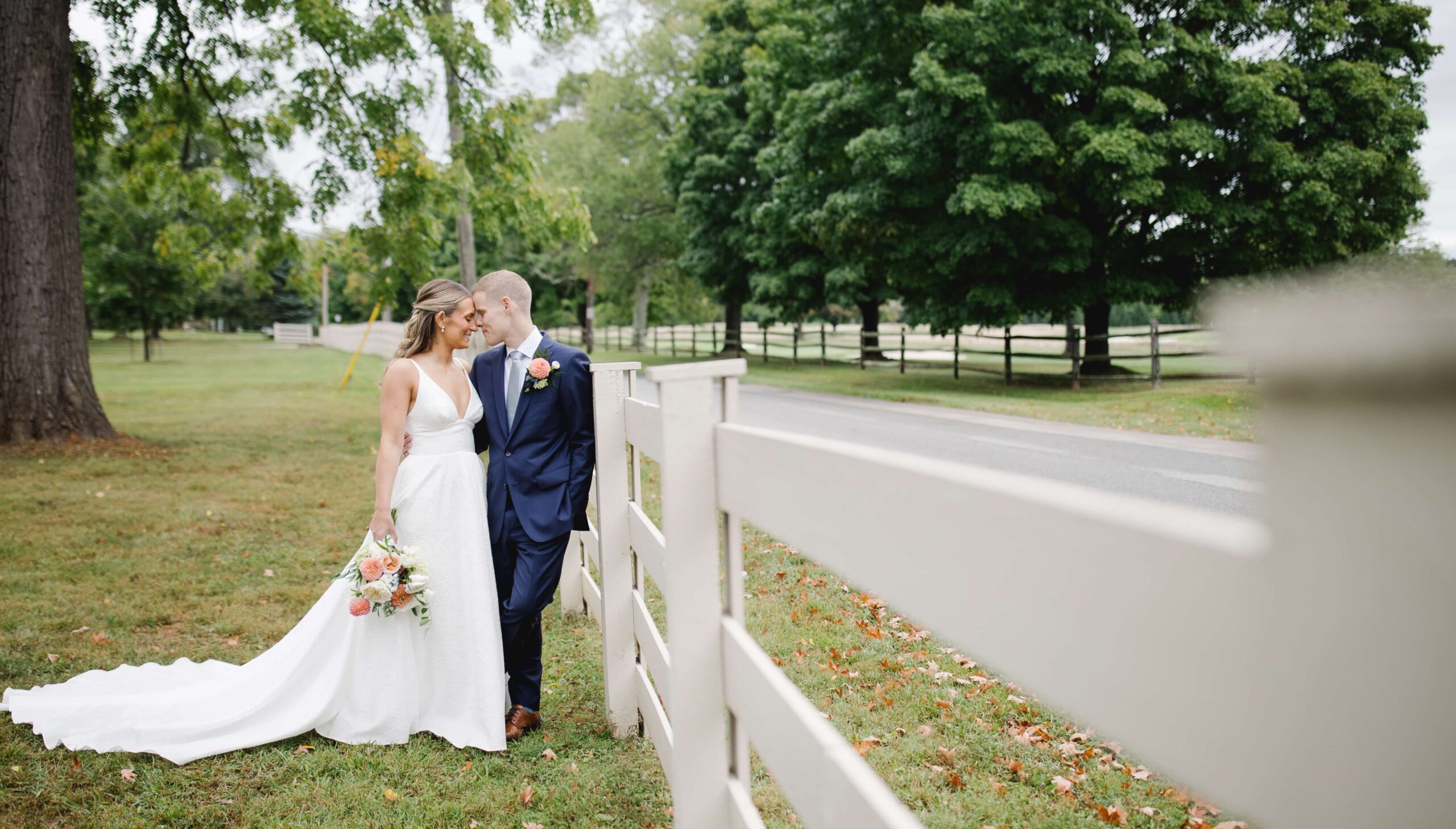 Bride and groom standing by a white fence in a park, embracing. The bride holds a bouquet, and trees are visible in the background.