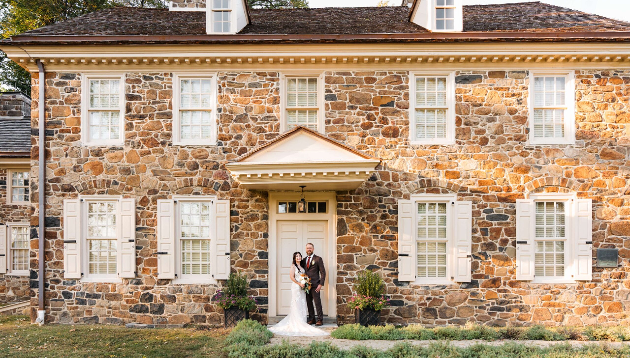 A couple in formal attire stands in front of a large stone house with white shutters and a wooden door.