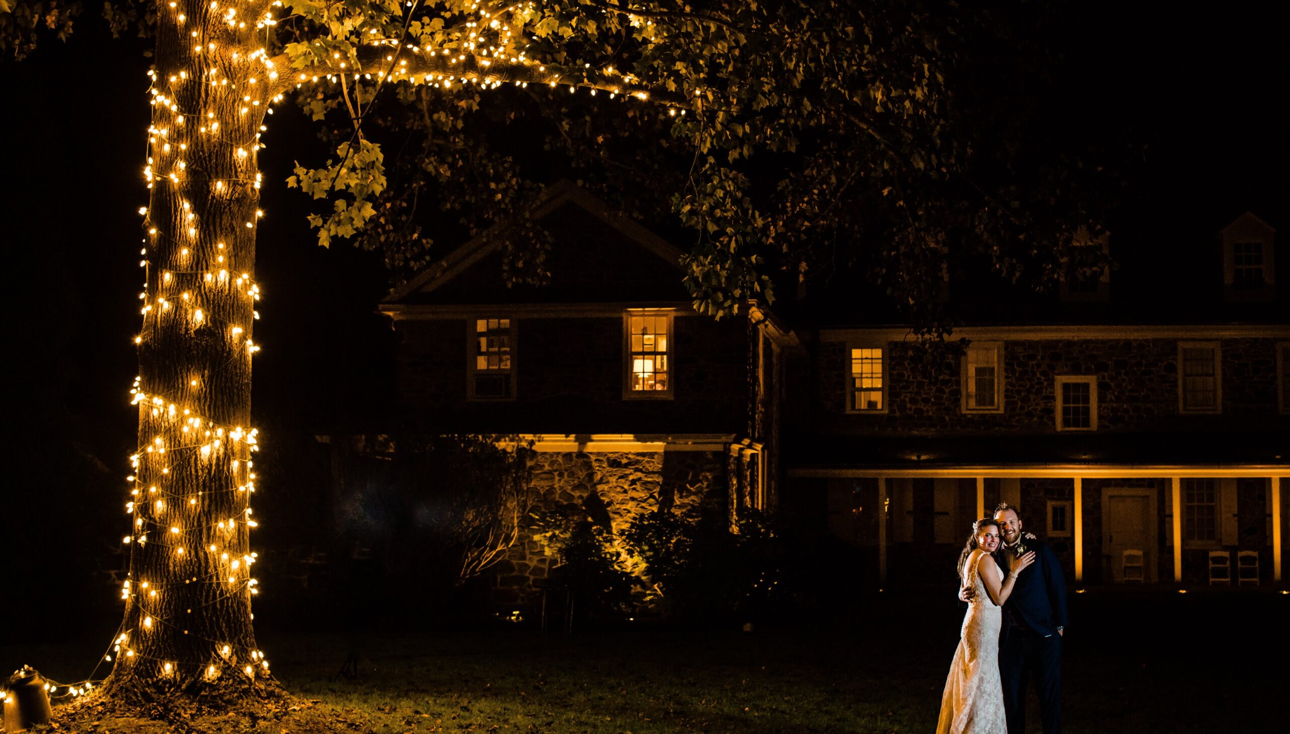 Bride and groom embrace under a tree wrapped in lights, in front of a large house at night.