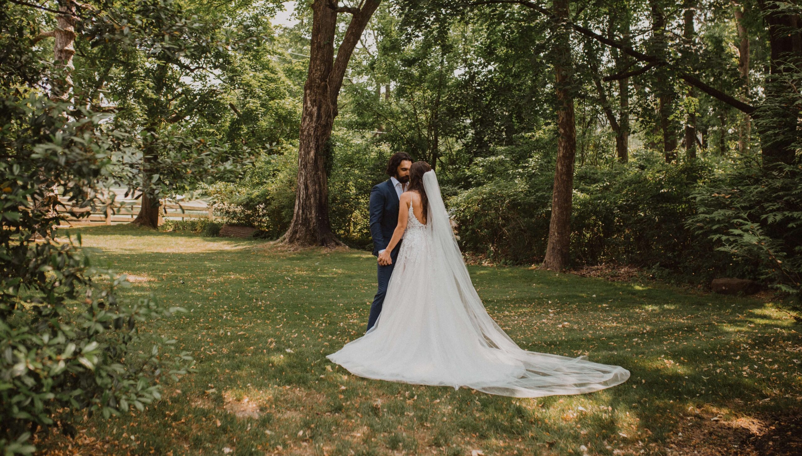 A bride and groom stand close together in a wooded area, with the bride in a white gown and long veil, and the groom in a navy suit, surrounded by green trees and grass.