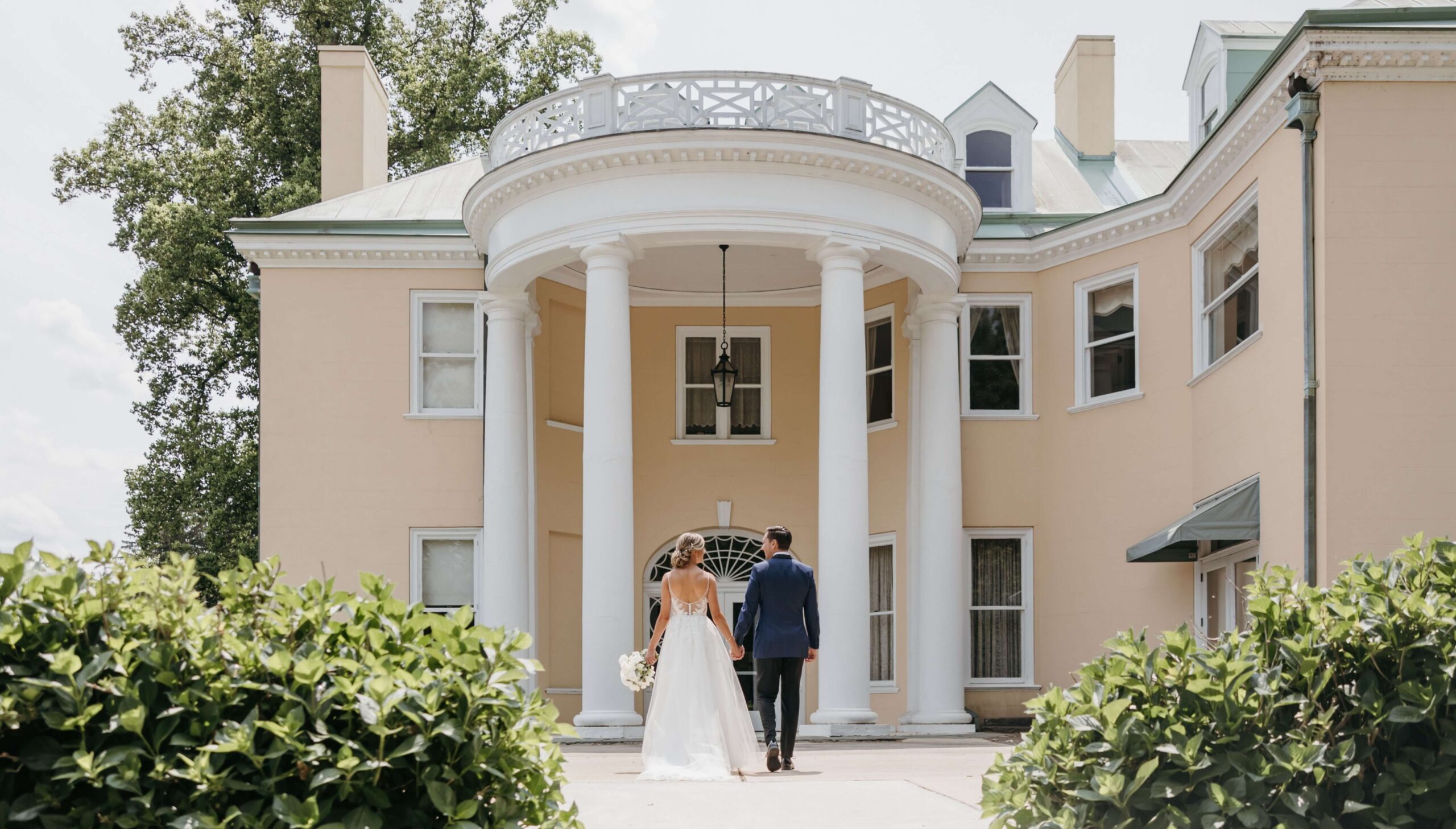 A bride and groom walk towards a large, elegant building with tall white columns and a curved balcony, surrounded by green bushes.