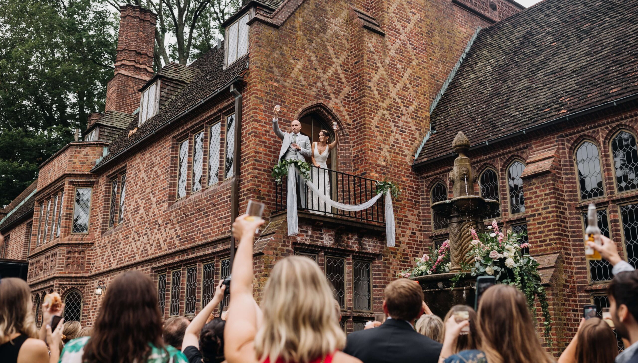 A couple stands on a balcony of a brick building, waving. Guests below raise glasses in a toast. A fountain decorated with flowers is in the foreground.