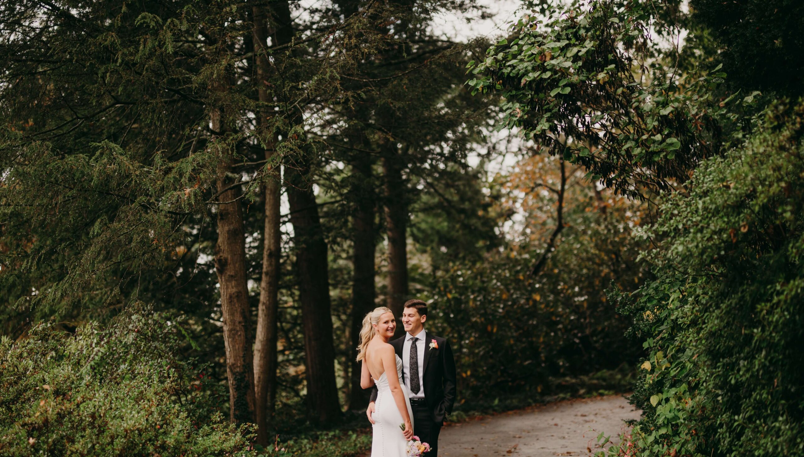 A bride and groom stand on a forest path surrounded by trees and greenery.