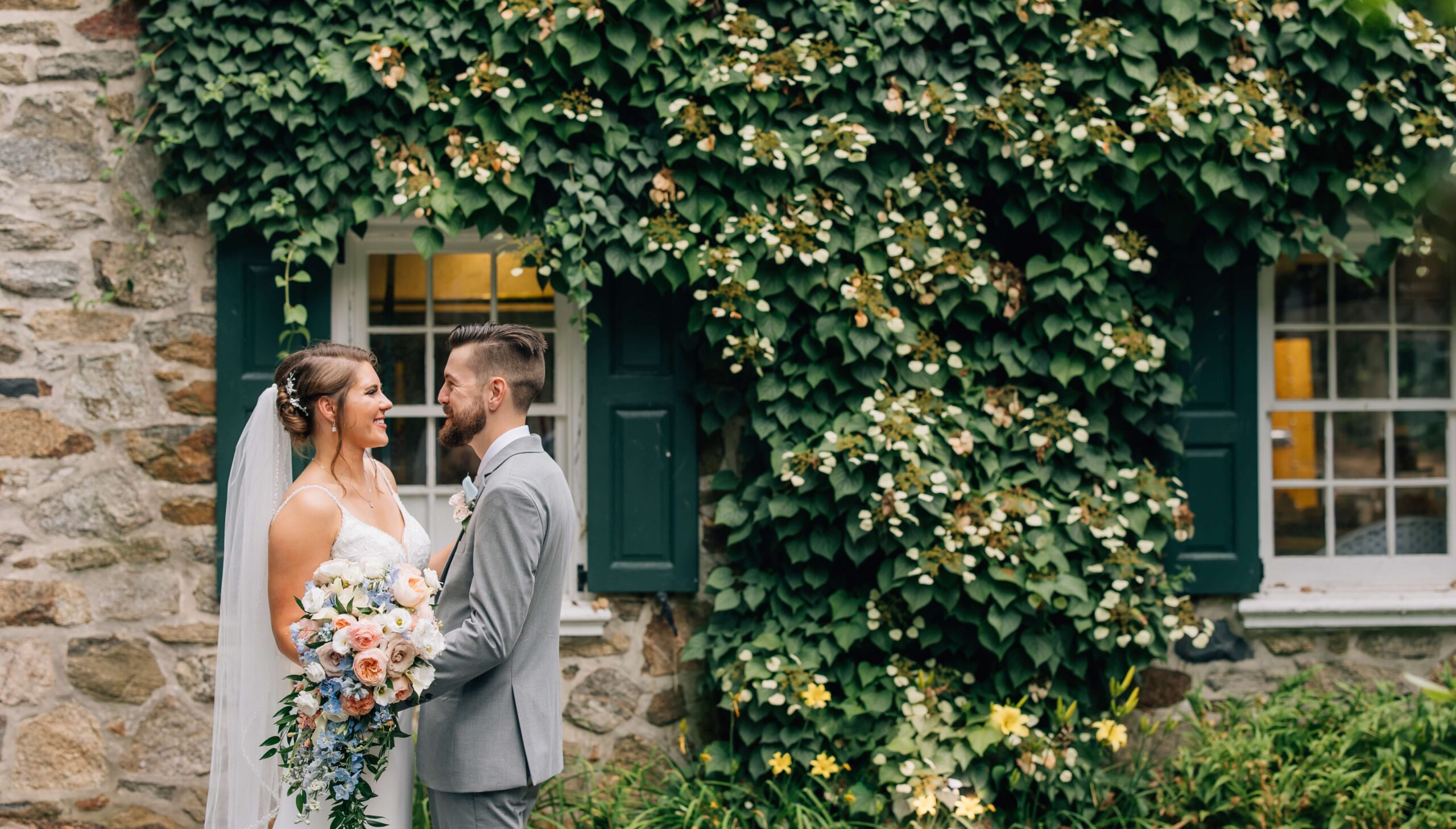 A bride and groom stand together holding a bouquet, smiling at each other in front of a stone house covered in green ivy.