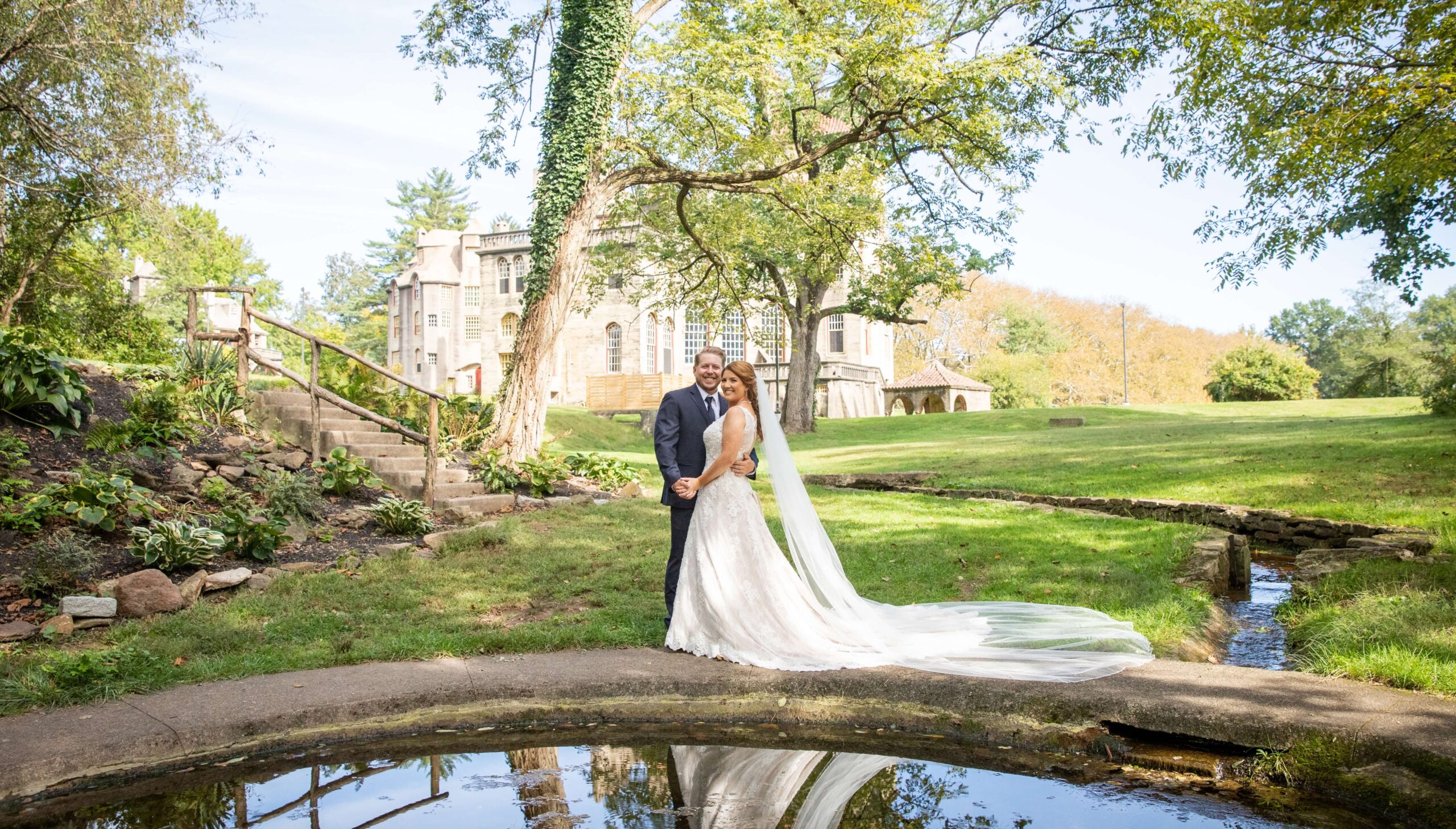 A bride and groom stand by a small pond, embracing, with a stone building and trees in the background.