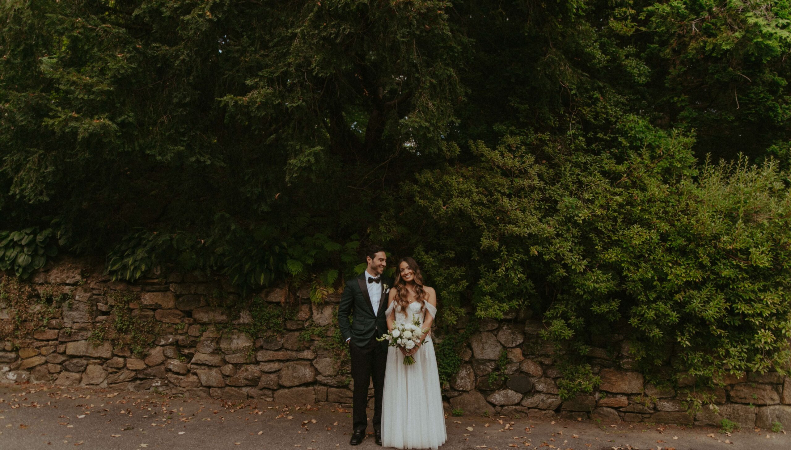 A bride and groom stand smiling in formal attire, holding a bouquet, in front of a stone wall and lush greenery.