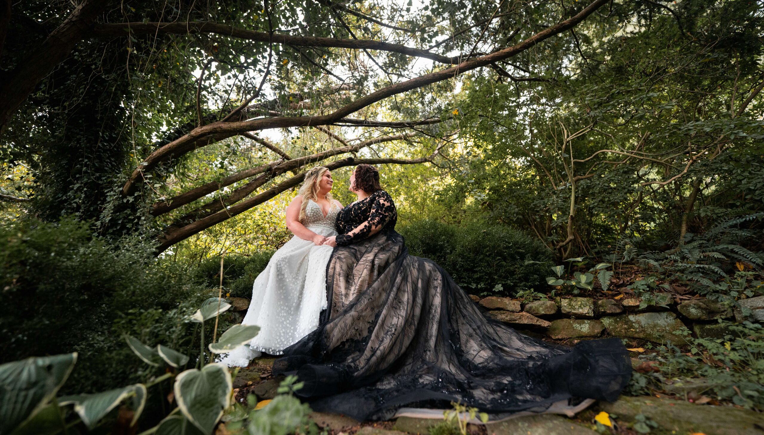 Two people in elegant gowns sit closely together in a lush garden setting. One gown is white, and the other is black with intricate details. Lush greenery surrounds them.