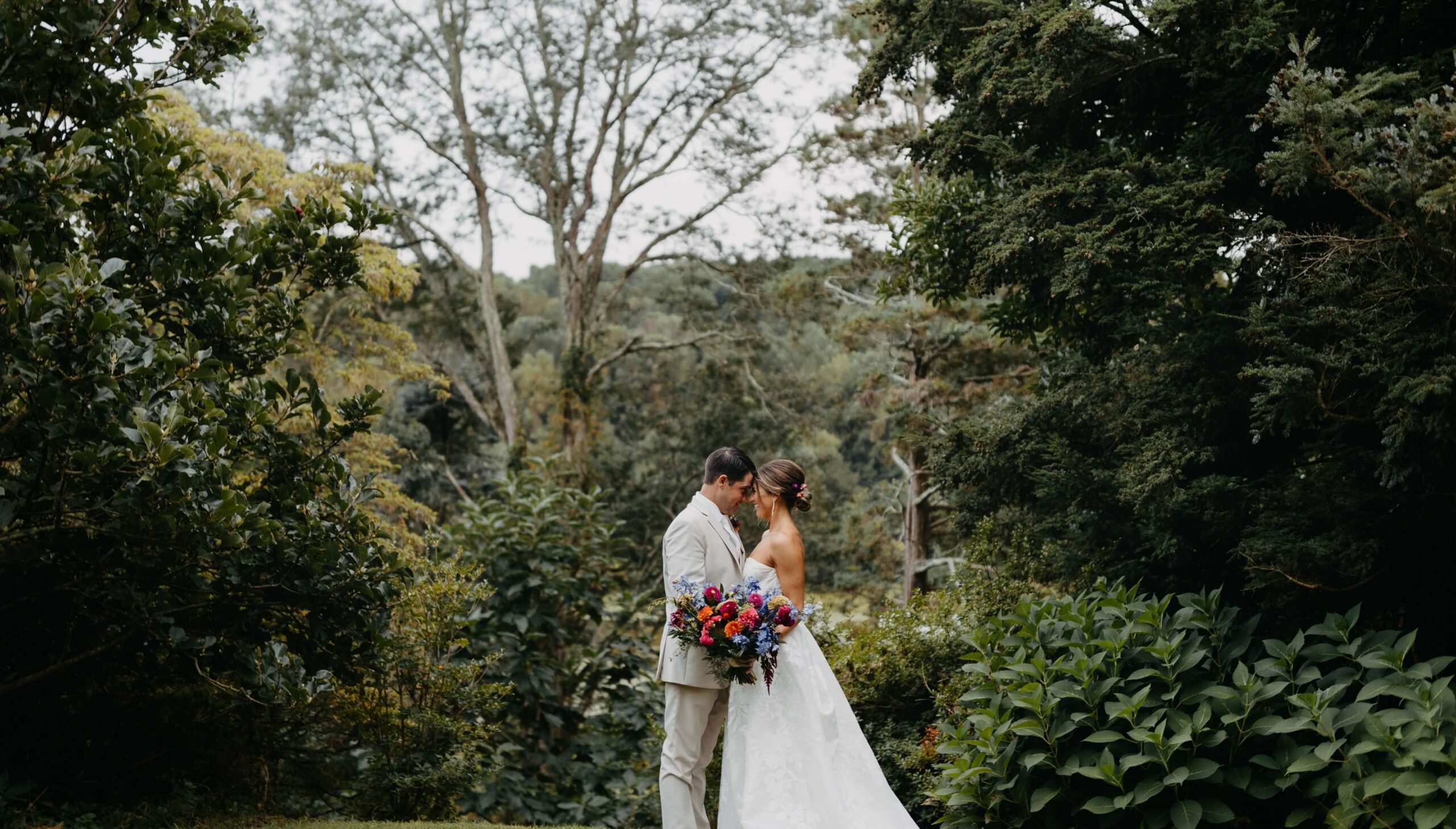 A bride and groom stand closely, gazing at each other, in a lush garden setting. The bride holds a colorful bouquet.