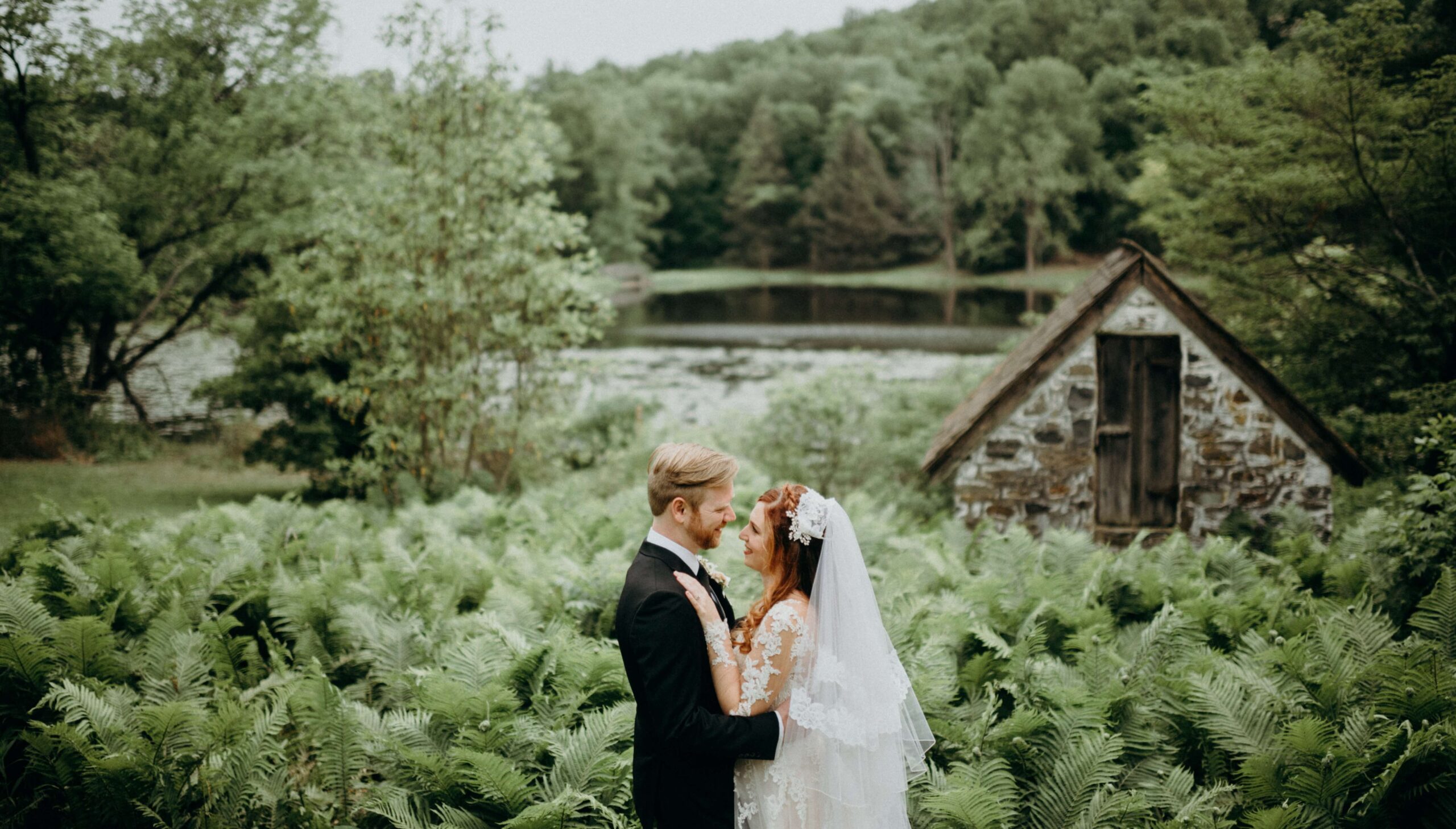 Bride and groom in wedding attire embrace in a lush, green garden with a stone cabin and pond in the background.