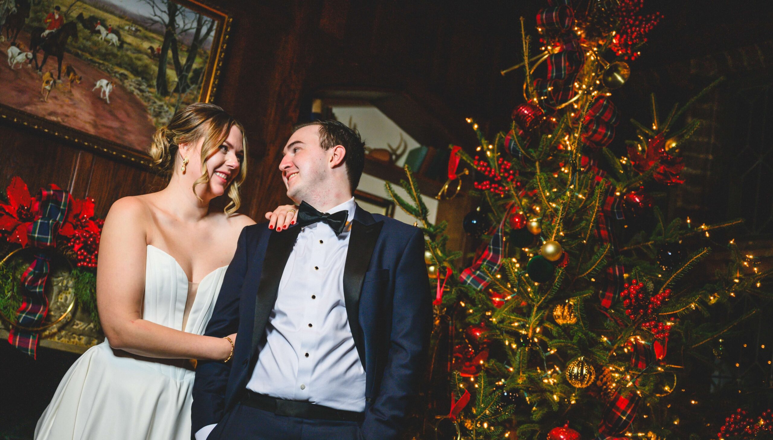 A couple in formal attire smiles at each other beside a decorated Christmas tree, with a painting and festive decorations in the background.