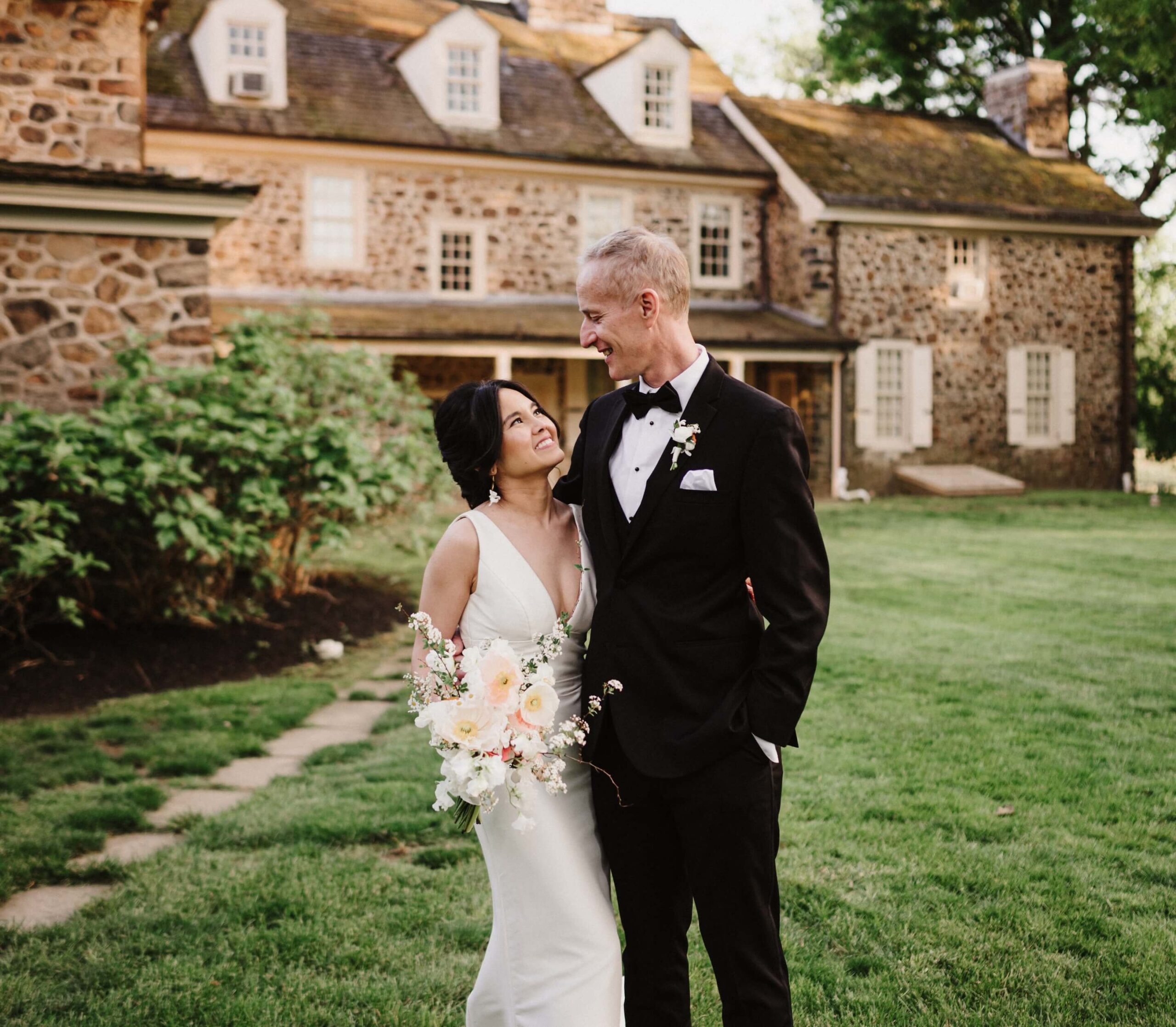 A bride in a white dress and groom in a black tuxedo smile at each other, standing on a grassy area in front of a stone building.