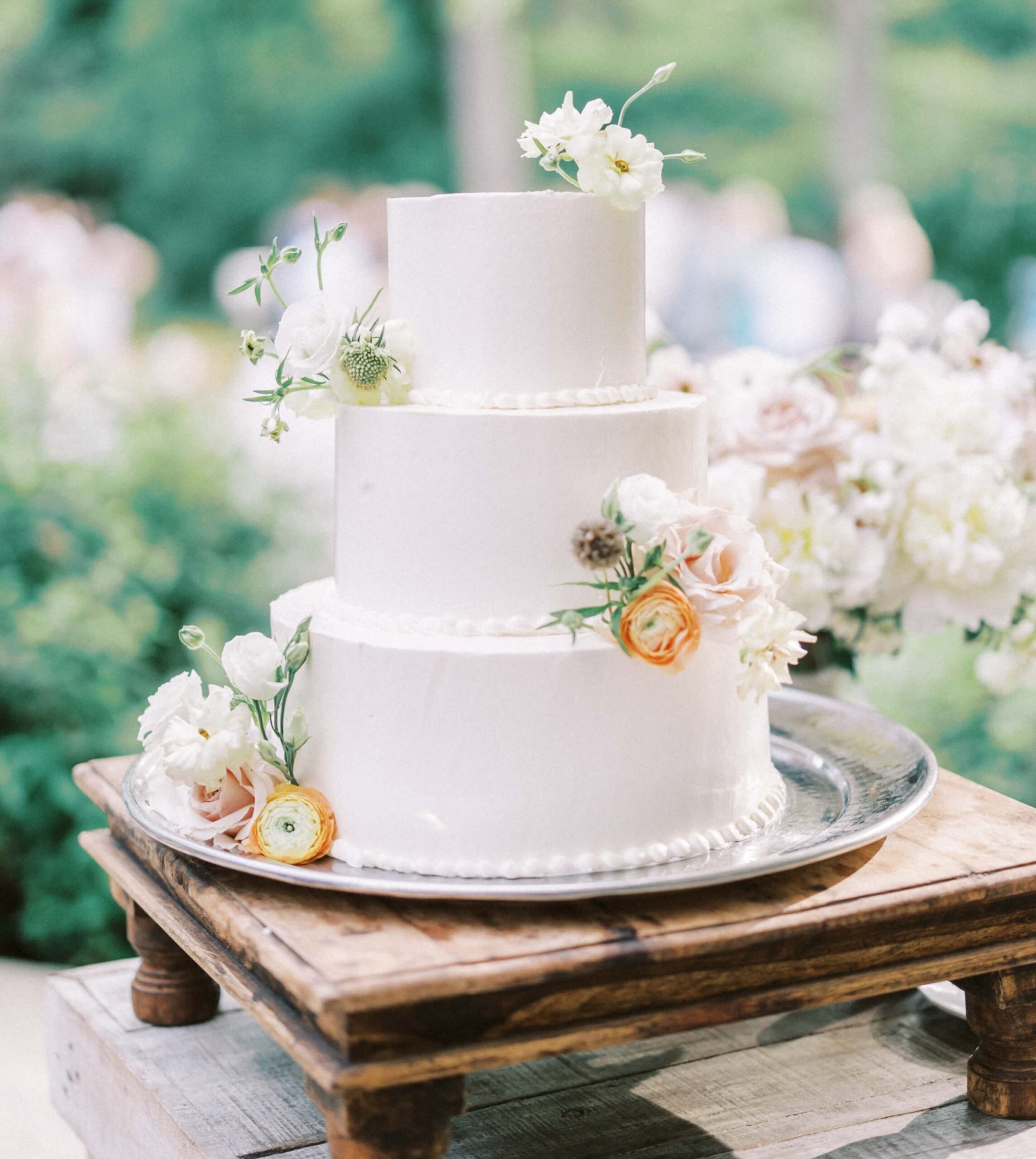 A three-tier white cake decorated with small flowers sits on a silver platter atop a wooden stand.