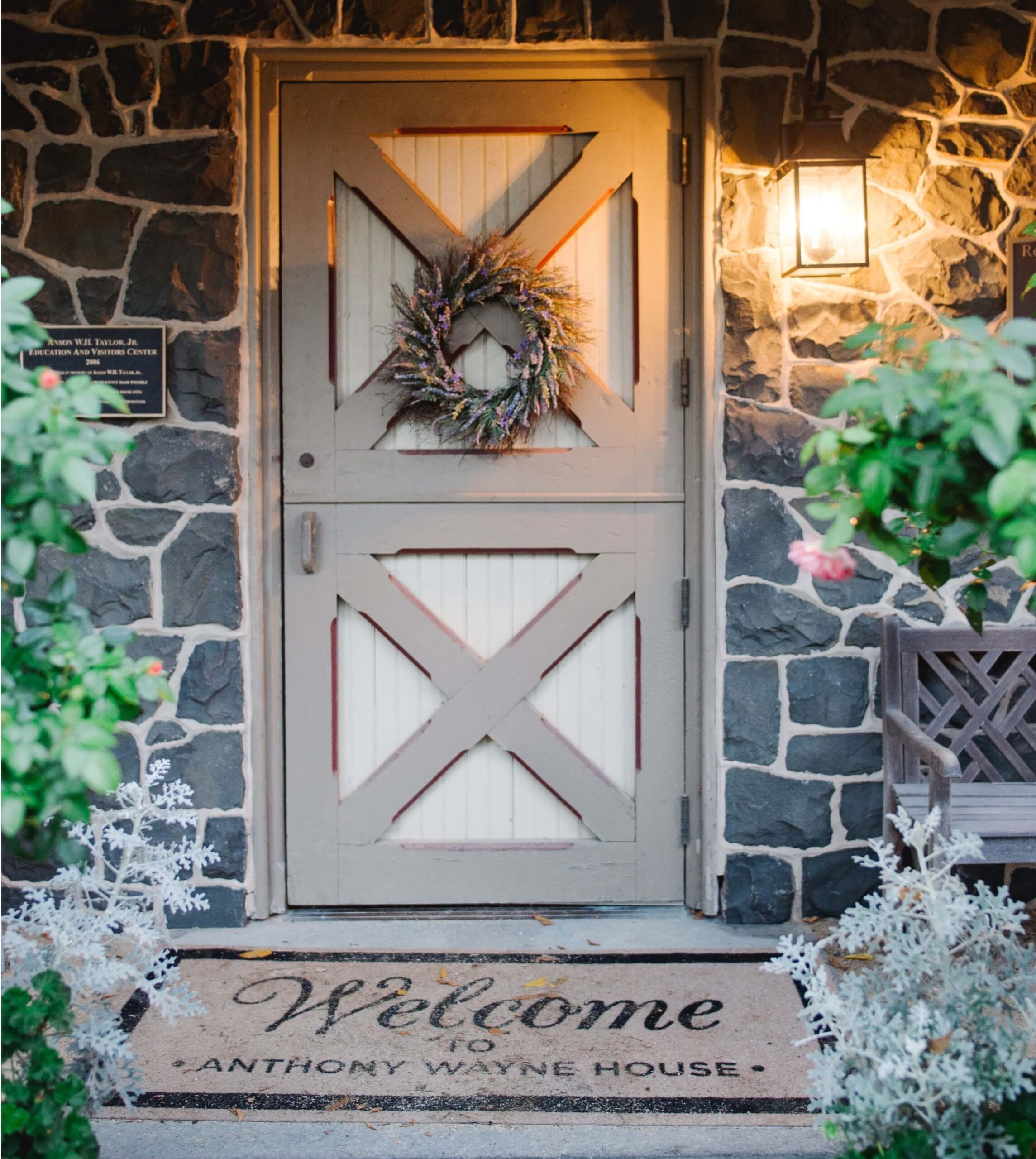 A rustic wooden door with a wreath, next to a lantern. A doormat reads