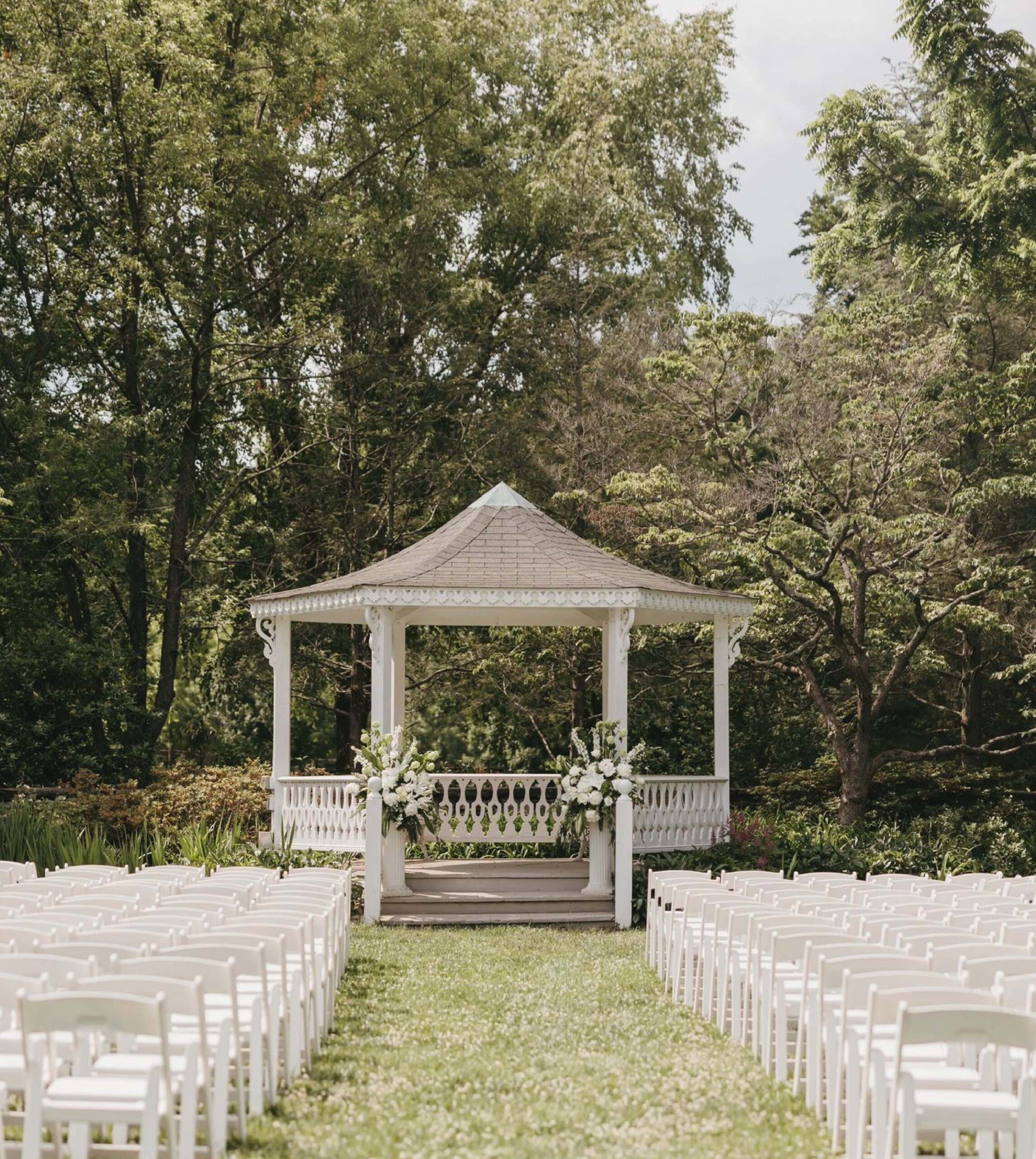 White gazebo in garden setting, surrounded by trees and greenery. Rows of white chairs lead to the gazebo, adorned with floral arrangements.
