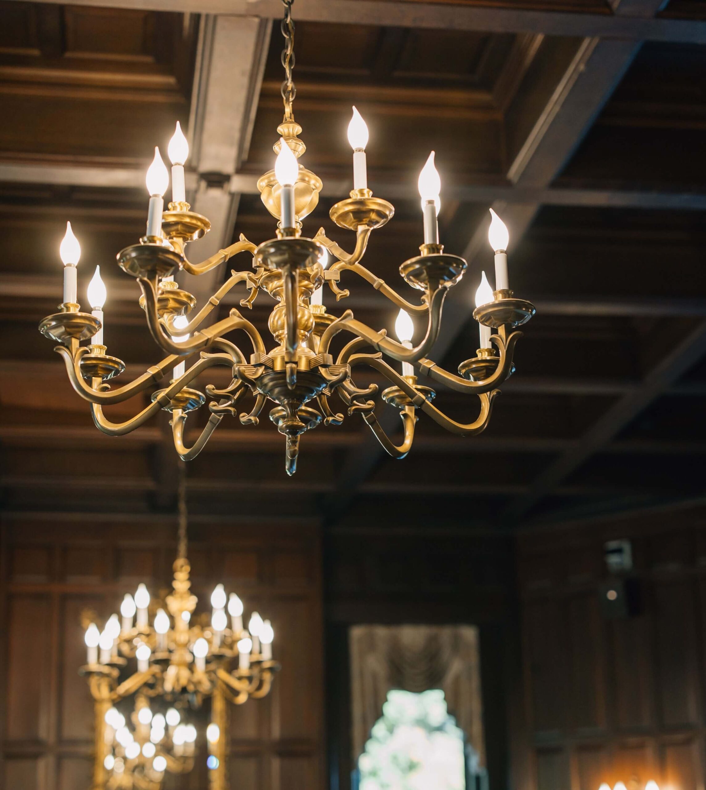 Ornate golden chandelier with lit candles hanging from a wooden ceiling, reflecting in a mirror below.