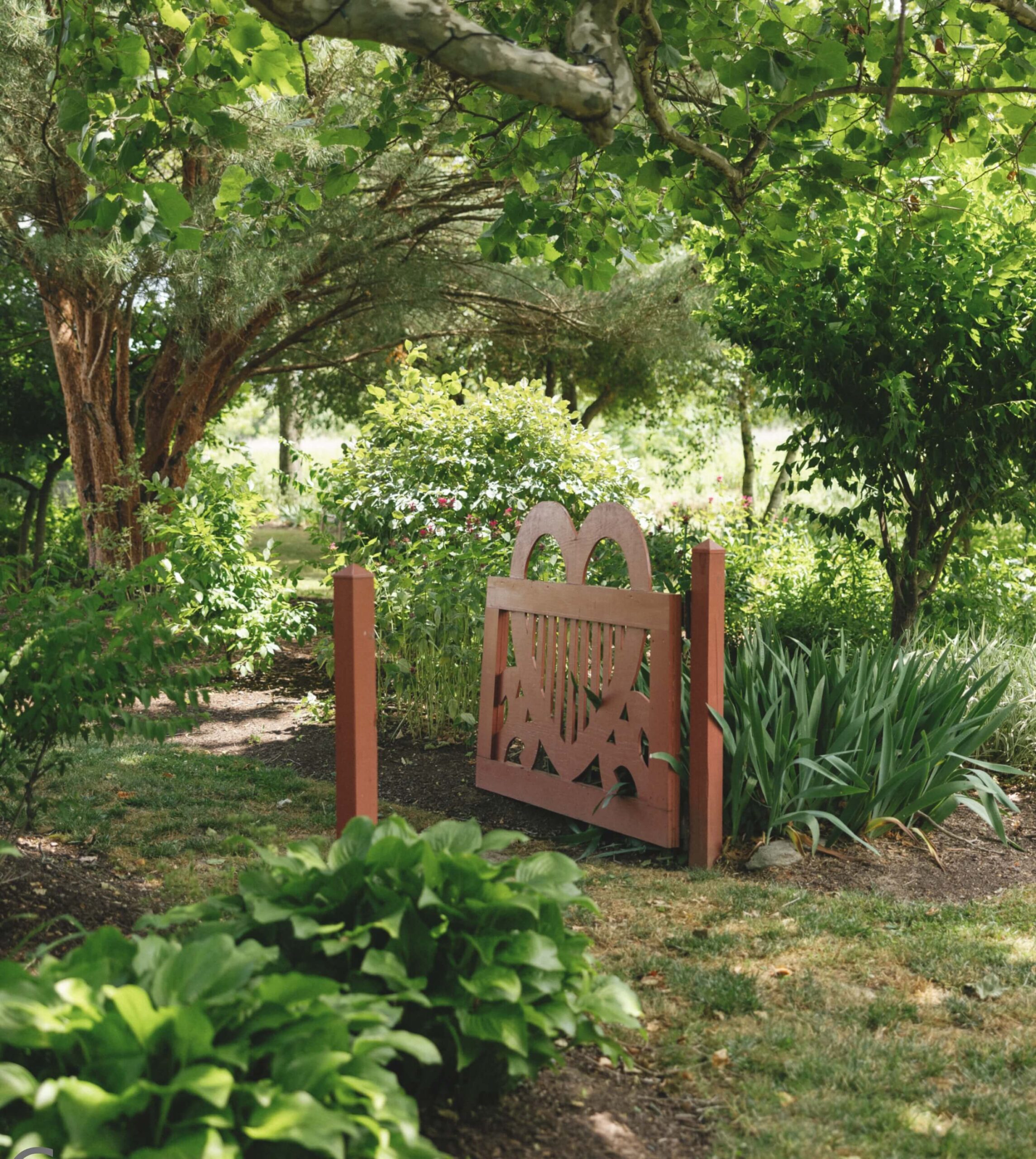 Wooden garden gate with heart-shaped designs, surrounded by lush green plants and trees.