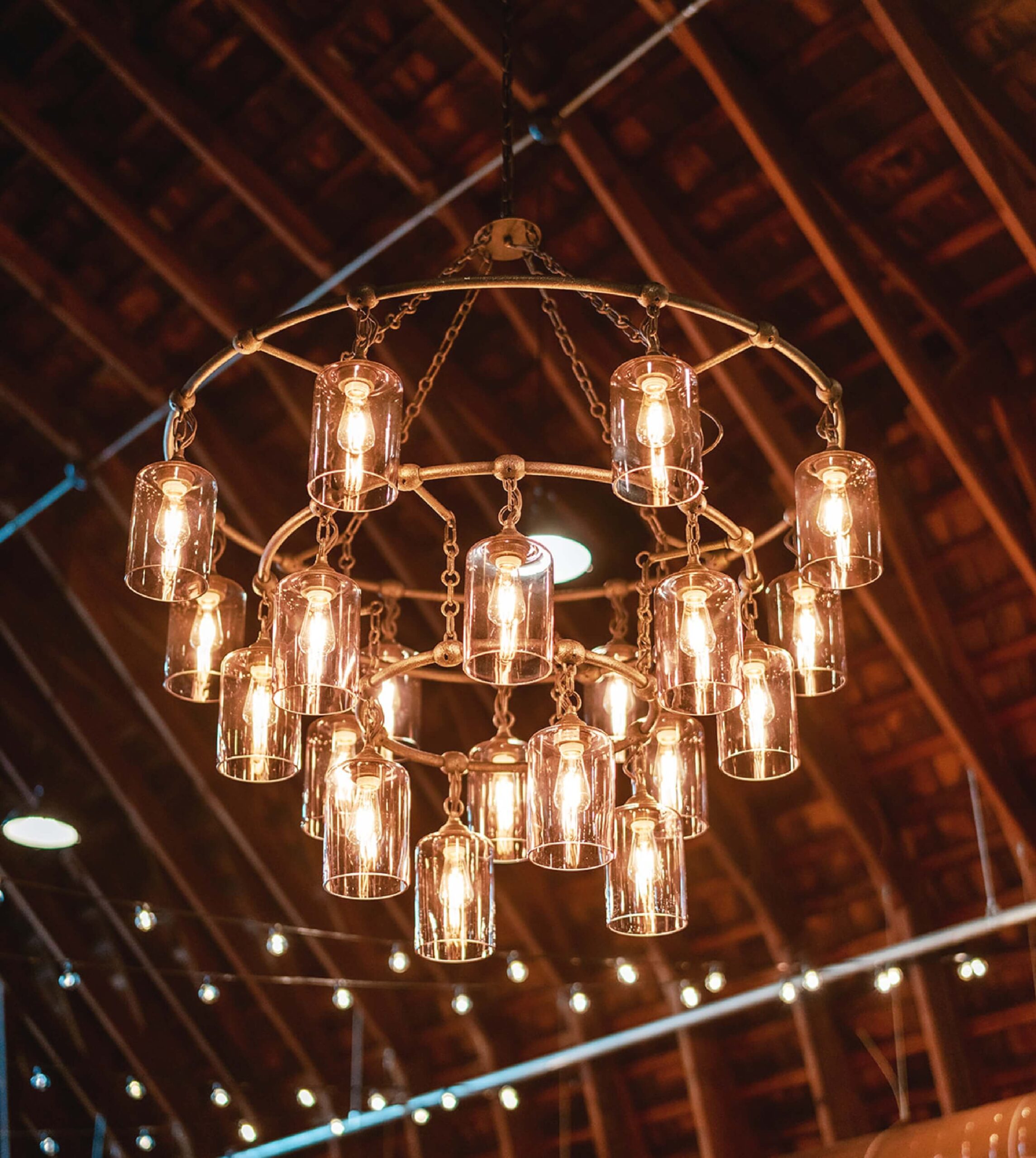 A metal chandelier with several exposed bulbs, each encased in glass, hangs in a wooden, barn-like ceiling. Strings of lights are visible in the background.