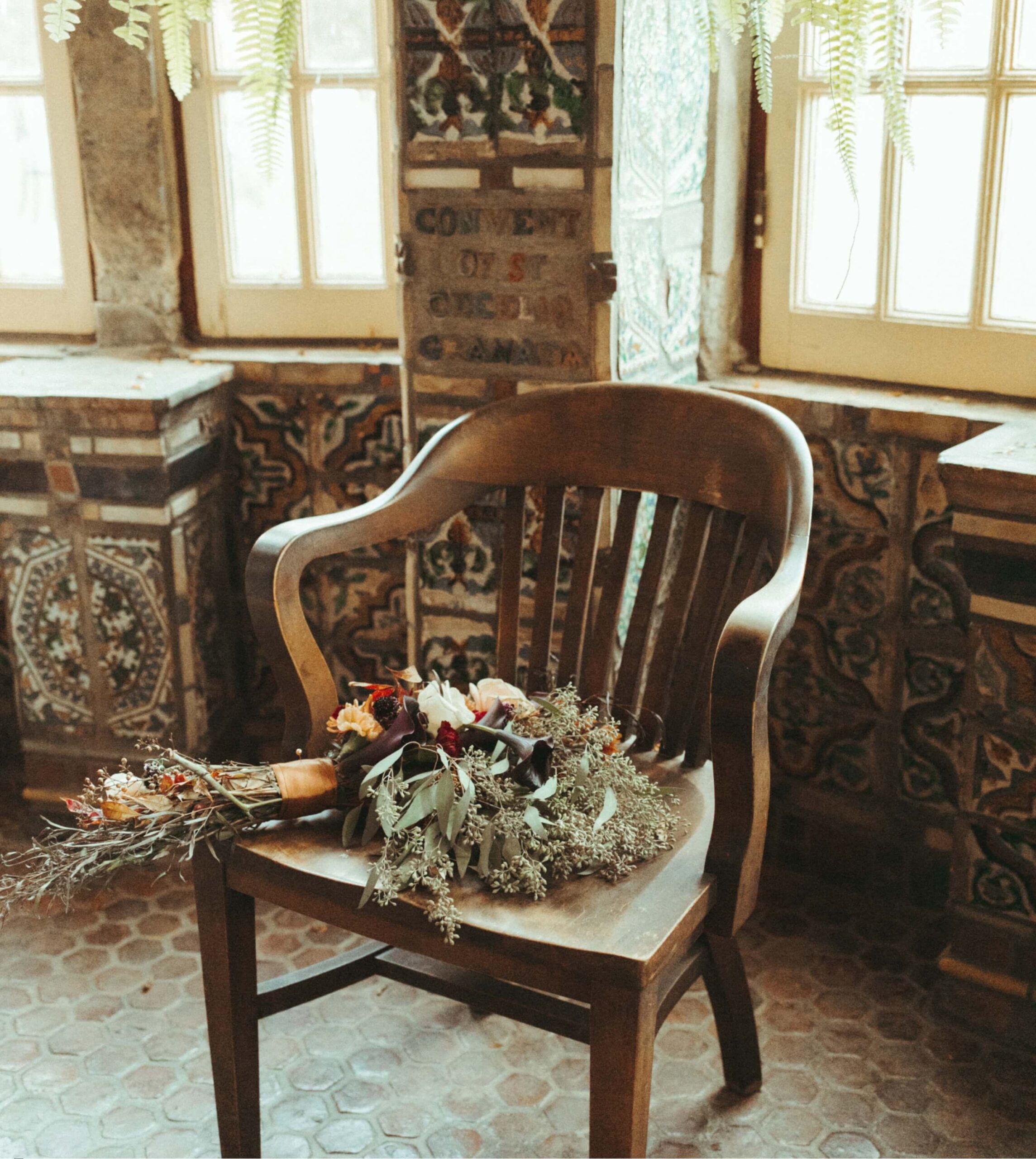 A wooden chair holds a bouquet of dried flowers in a room with patterned tiles and large windows.