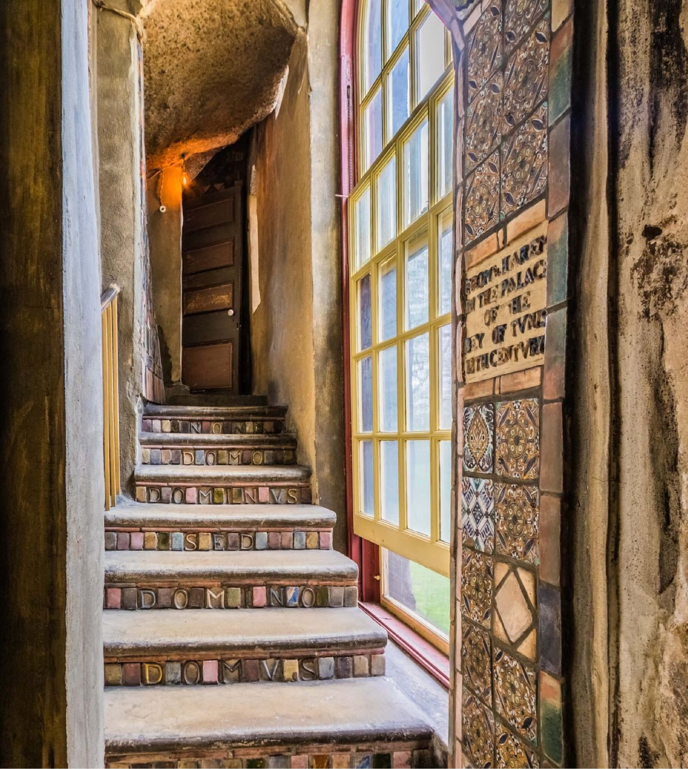 Stone staircase with decorative text on the risers, beside a large window and a tiled wall, leading to a wooden door at the top.